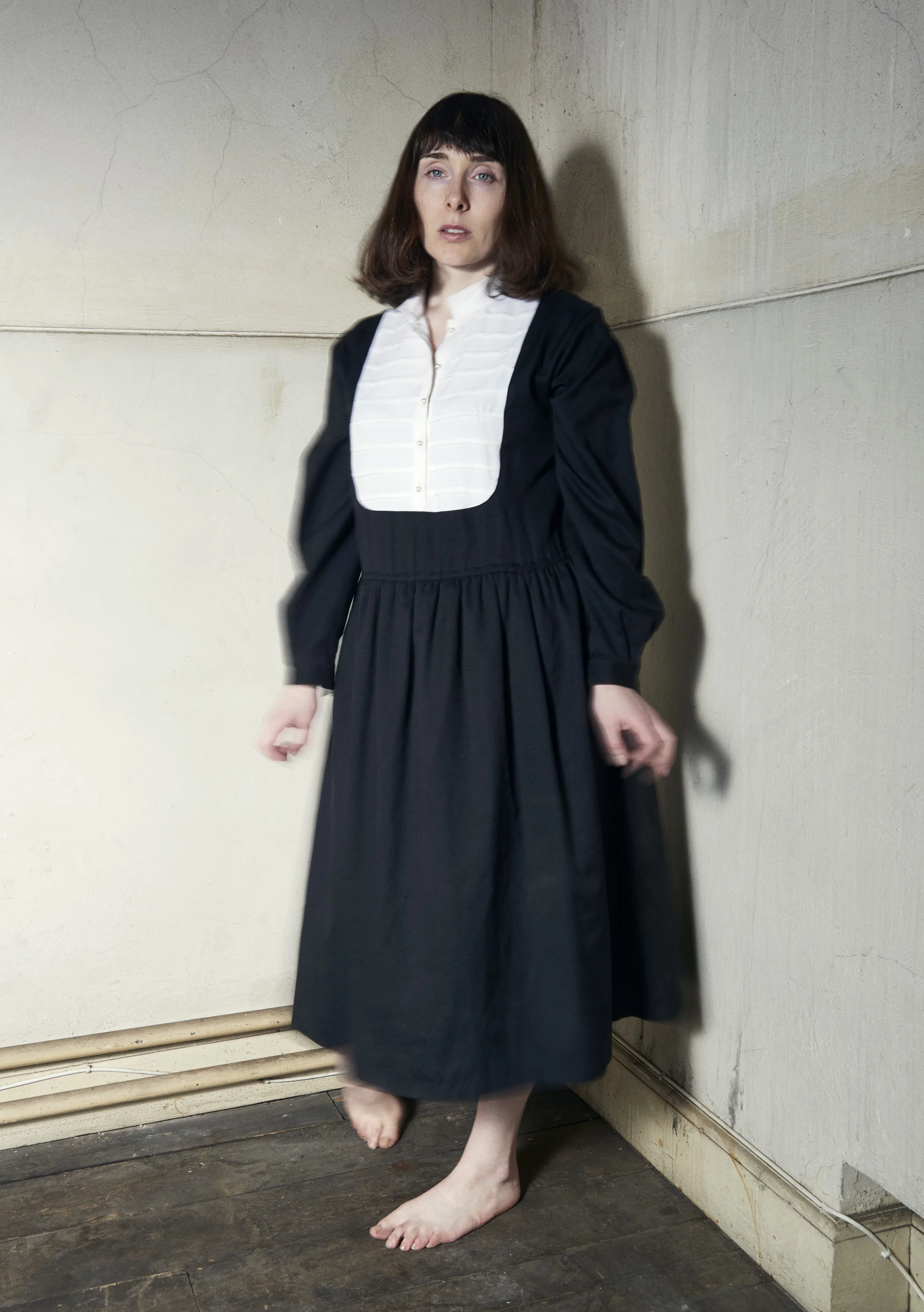 A woman with shoulder-length brown hair, pale skin, and makeup standing barefoot against a plain, textured wall. She wears a long, dark dress with a white, buttoned-up top. She is looking slightly upward with a neutral expression, and her left hand h