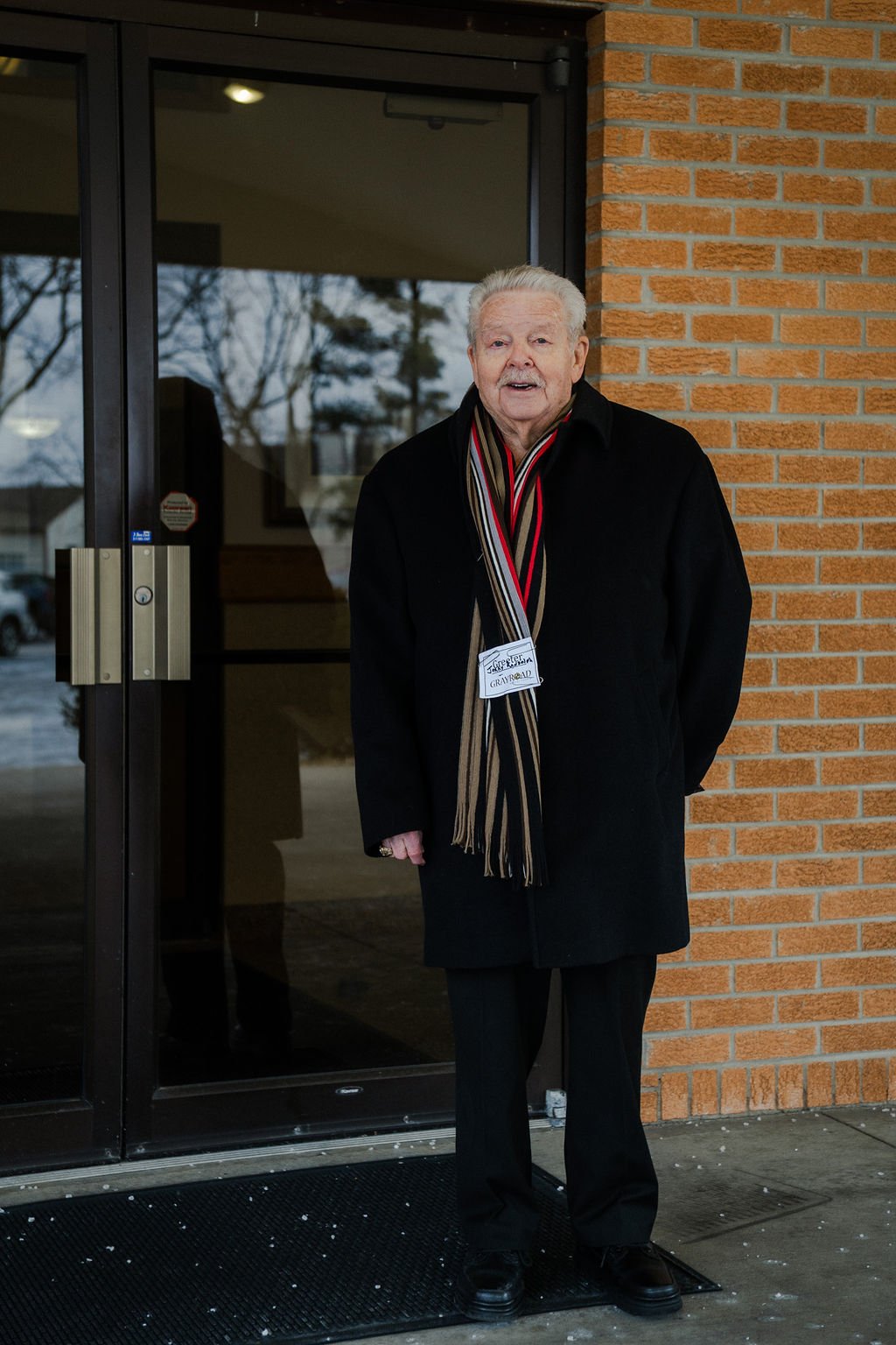 Older man with light hair, standing outside a building near a glass door, wearing a black coat and a striped scarf, with a name tag that reads "GREETER" and "GRAYBEARD."