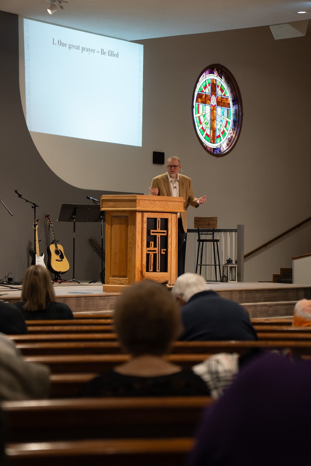 A man giving a speech at a church service or event, standing at a wooden pulpit with a cross design, with an audience seated in pews. Behind him, there are guitars, a music stand, and a large stained glass window depicting a cross.
