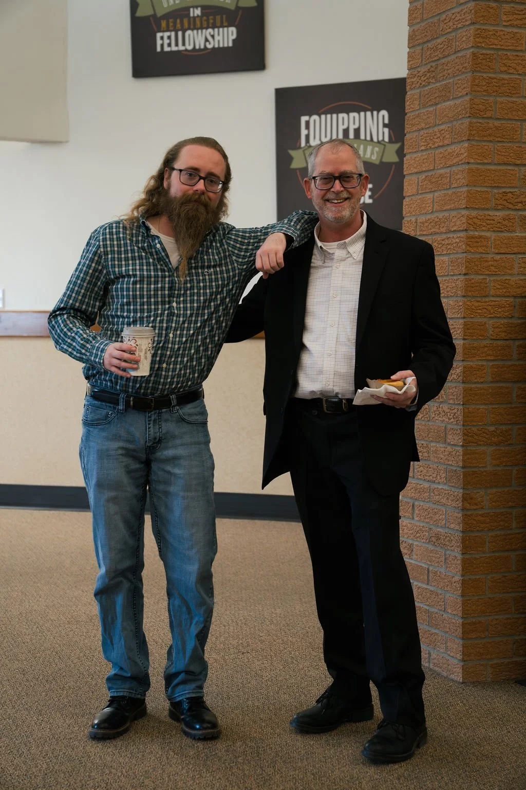 Two men standing together, one with long hair and beard holding a coffee cup, the other with short hair and glasses holding food, smiling in an indoor setting with signs on the wall.
