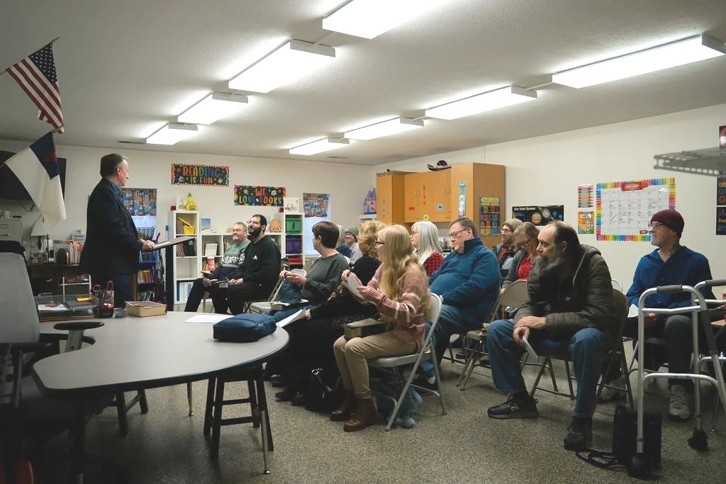 A man in a suit speaking to a group of adults seated in a classroom or community room with educational posters, bookshelves, and cabinets.