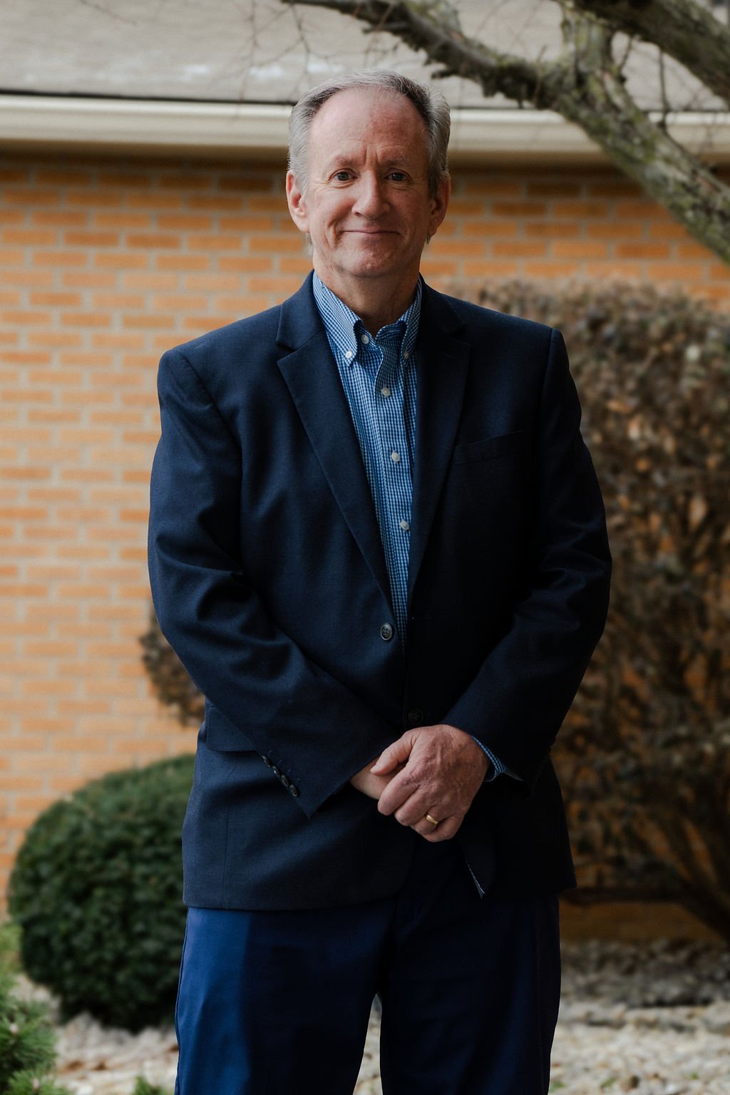 A middle-aged man standing outdoors in front of a brick wall and shrubbery, wearing a dark suit and blue checkered shirt, smiling slightly.