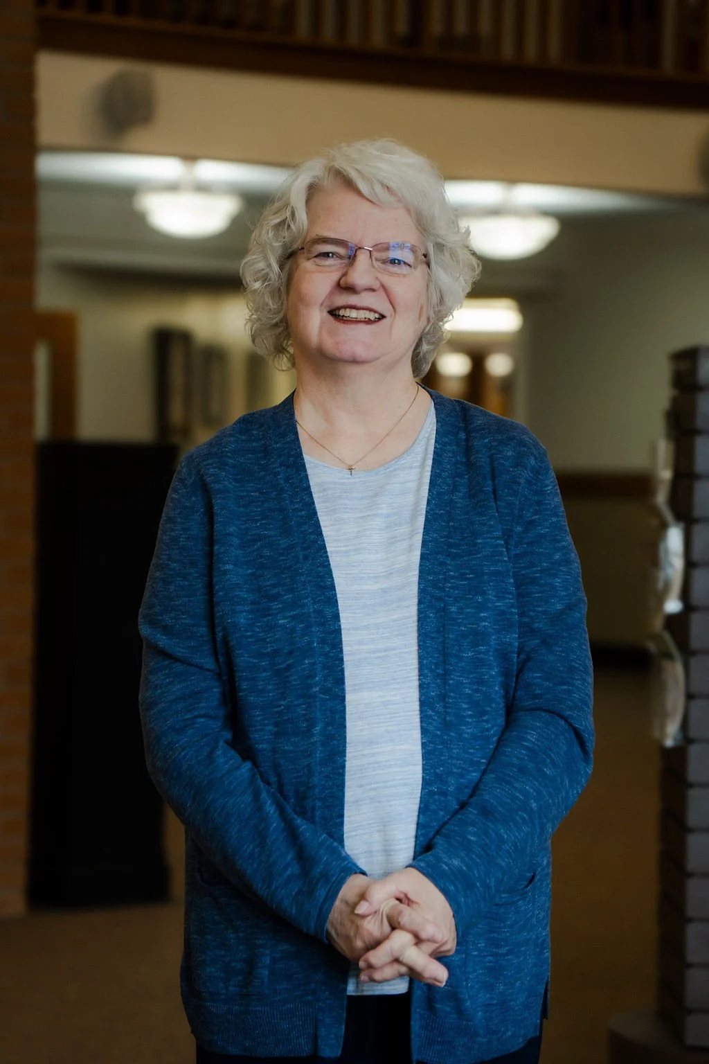 Smiling elderly woman with curly gray hair, glasses, and a blue cardigan stands indoors with a library in the background.