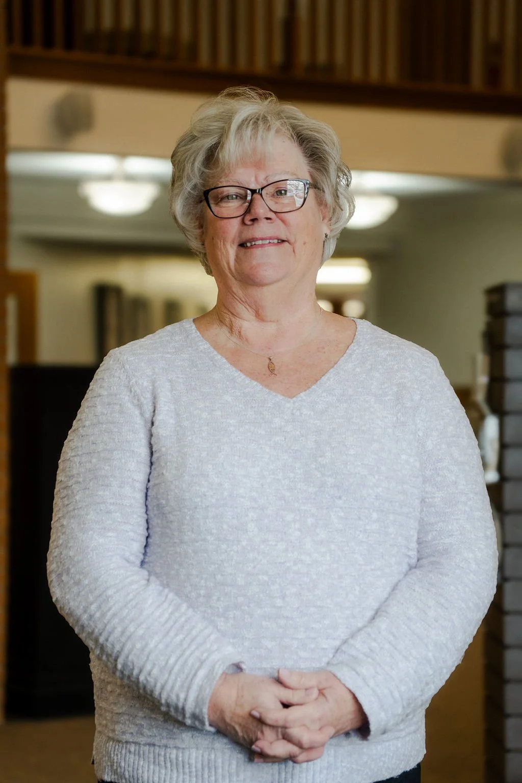 An older woman with short gray hair, glasses, and a light-colored sweater standing indoors and smiling.