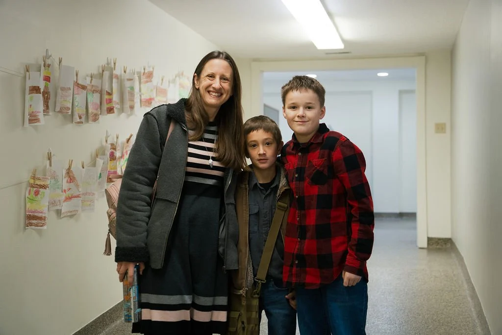 A woman and two boys smiling and standing in a hallway decorated with children's artwork on the wall.