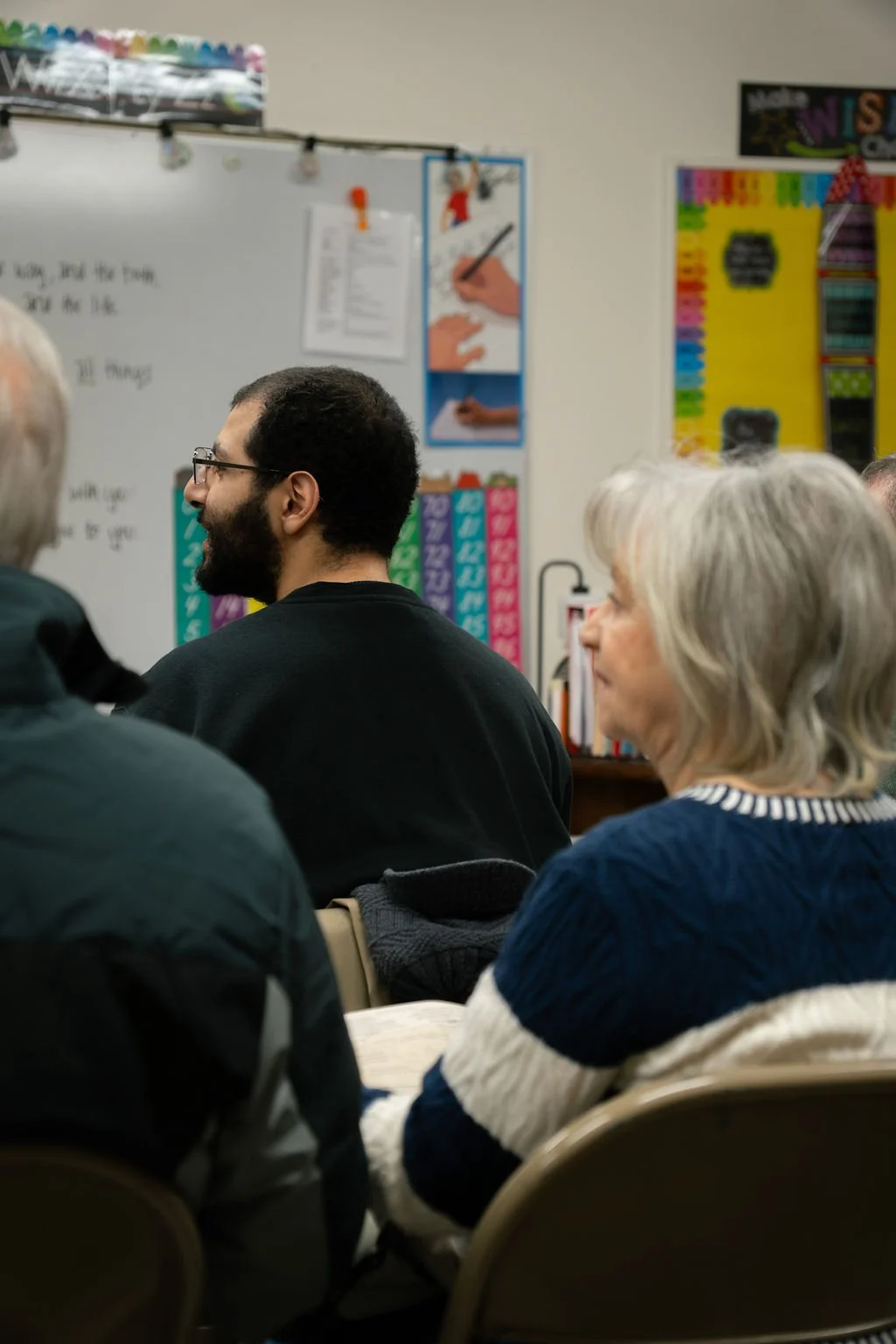 A group of people attending a class or meeting, seated and facing a whiteboard and classroom decorations in a school classroom.