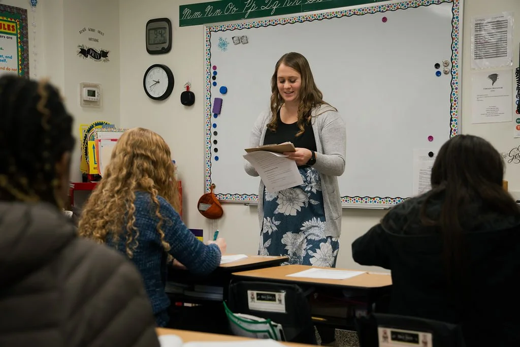 A teacher standing at the front of a classroom with students sitting at their desks, holding a clipboard and smiling. The classroom has a whiteboard, clocks, and educational posters on the walls.