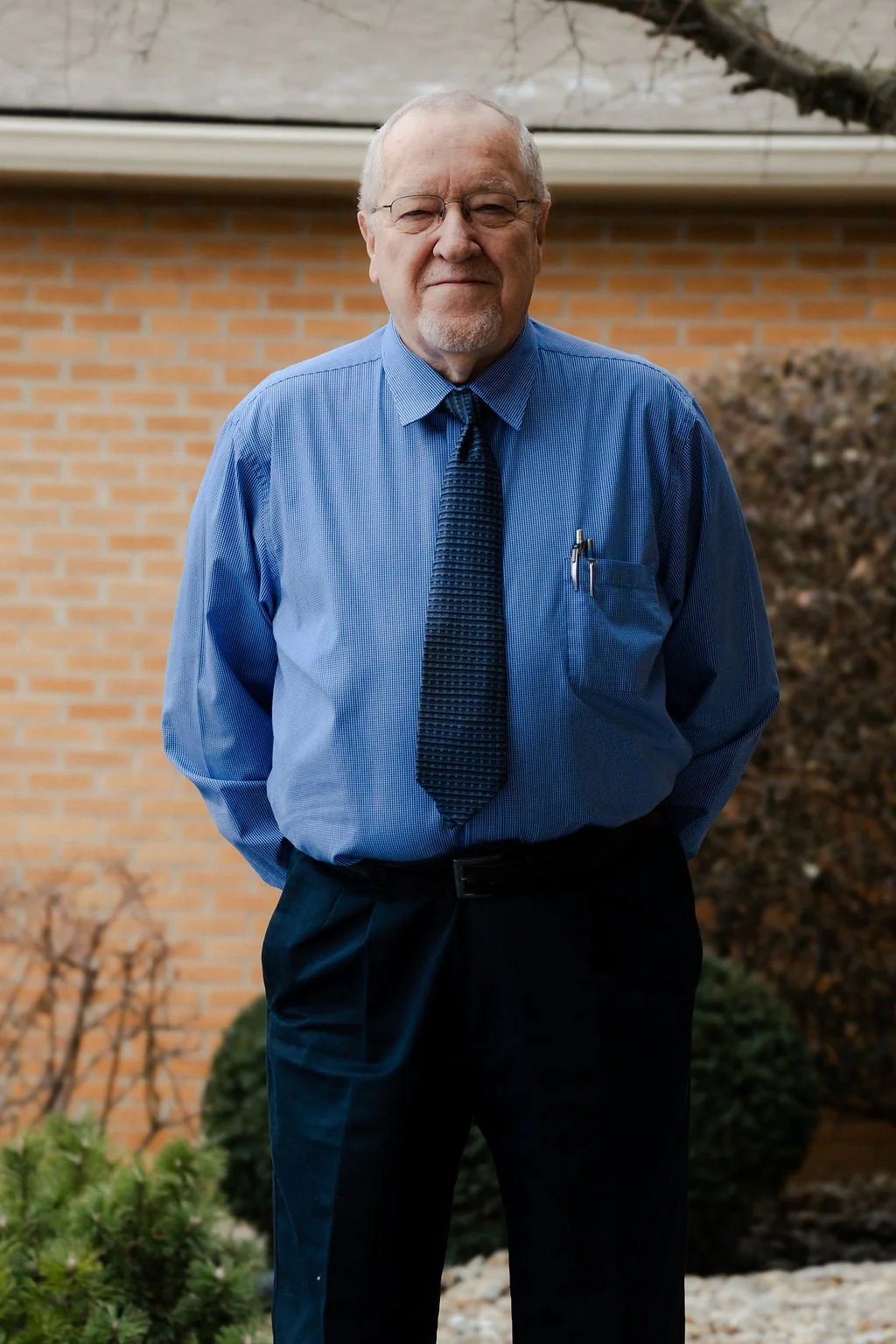 An elderly man with glasses, a light beard, and short gray hair standing outdoors in front of a brick wall, wearing a blue dress shirt, a patterned tie, and black pants.
