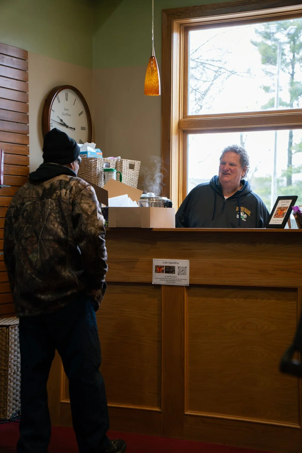 A man wearing a camouflage jacket and black beanie at a counter talking to a smiling man wearing a dark hoodie inside a cafe with a large window.