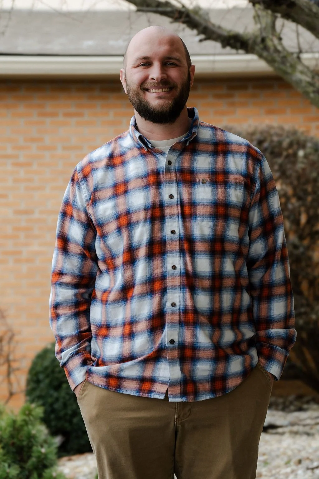 A smiling man with a beard, wearing a plaid shirt and khaki pants, standing outdoors in front of a brick house and a tree.