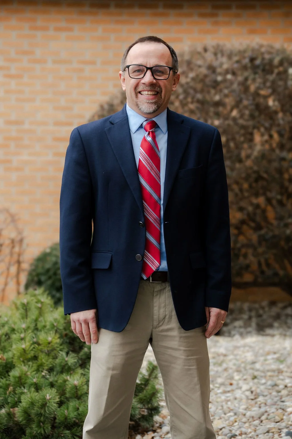A man wearing glasses, a navy blazer, a light blue shirt, a red and white striped tie, and beige pants, standing outdoors in front of a brick wall and bushes.