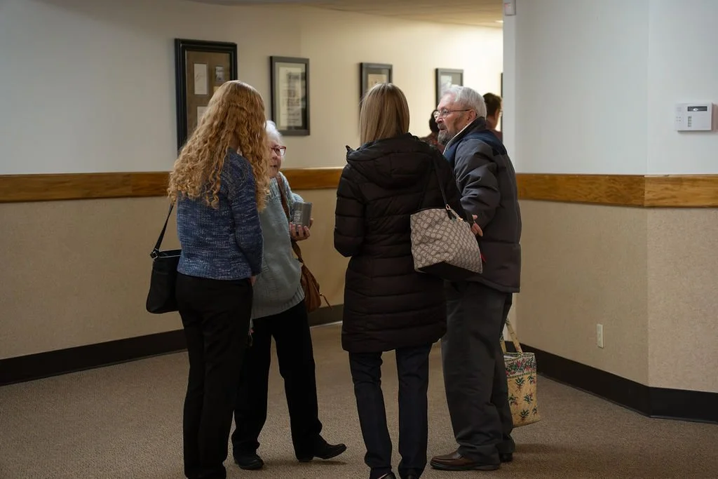Group of four adults engaged in conversation in a hallway, with framed pictures on the wall behind them.