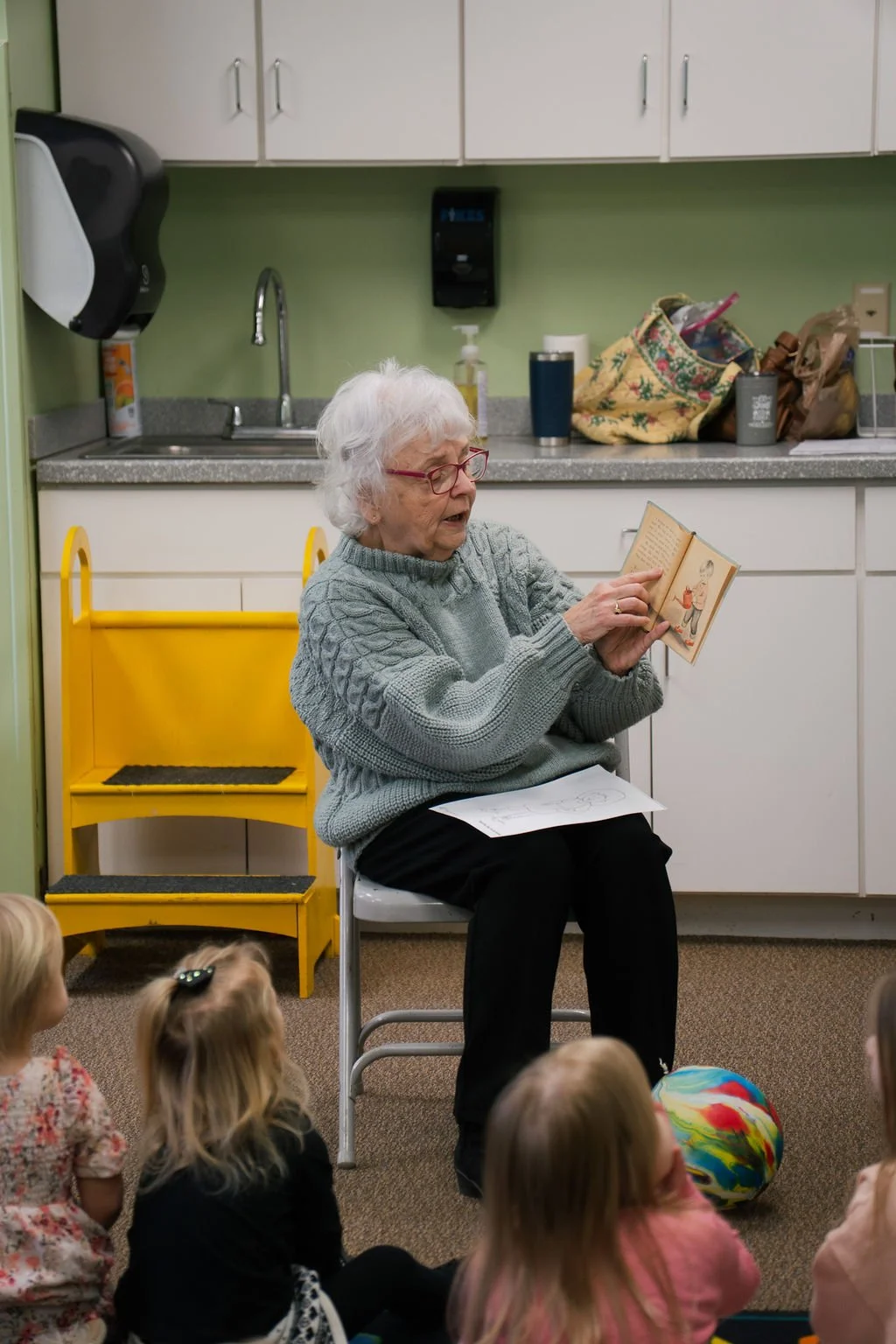 An elderly woman with white hair and glasses reading a book to young children sitting on the floor in a room with a yellow step stool and kitchen background.
