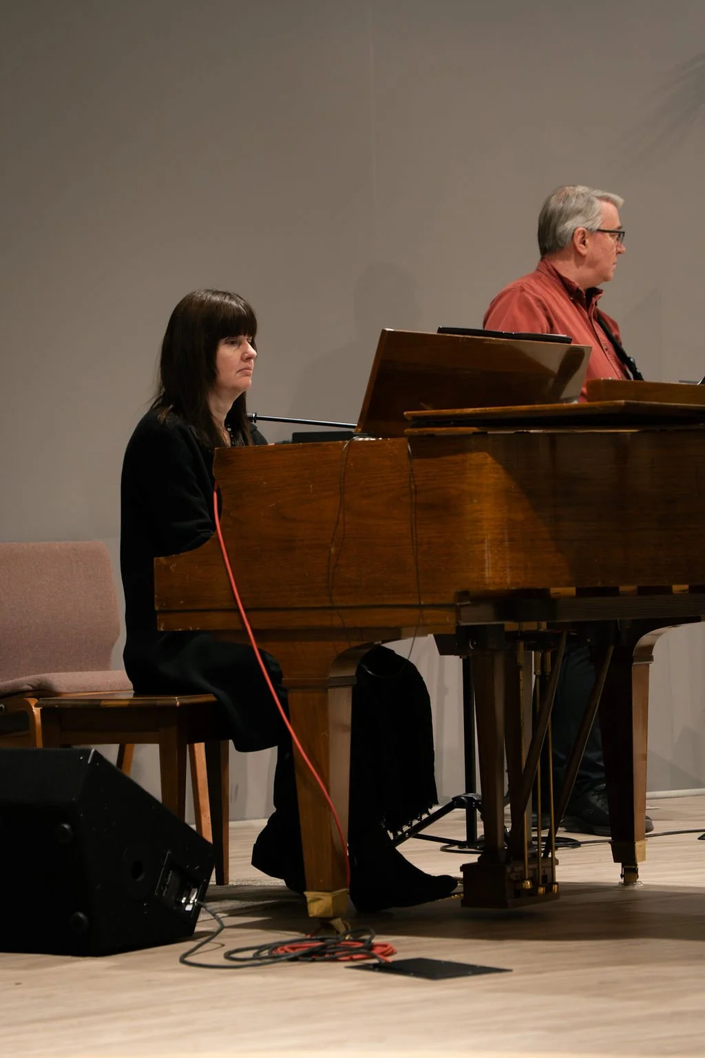 A woman playing a grand piano on stage, with a man standing nearby, both facing away from the camera.