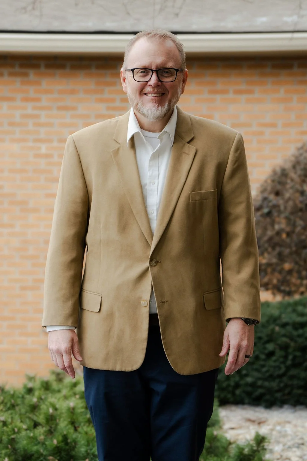 A man with glasses, a white beard, wearing a beige blazer, white shirt, and dark pants standing outdoors with a brick building and bushes in the background.