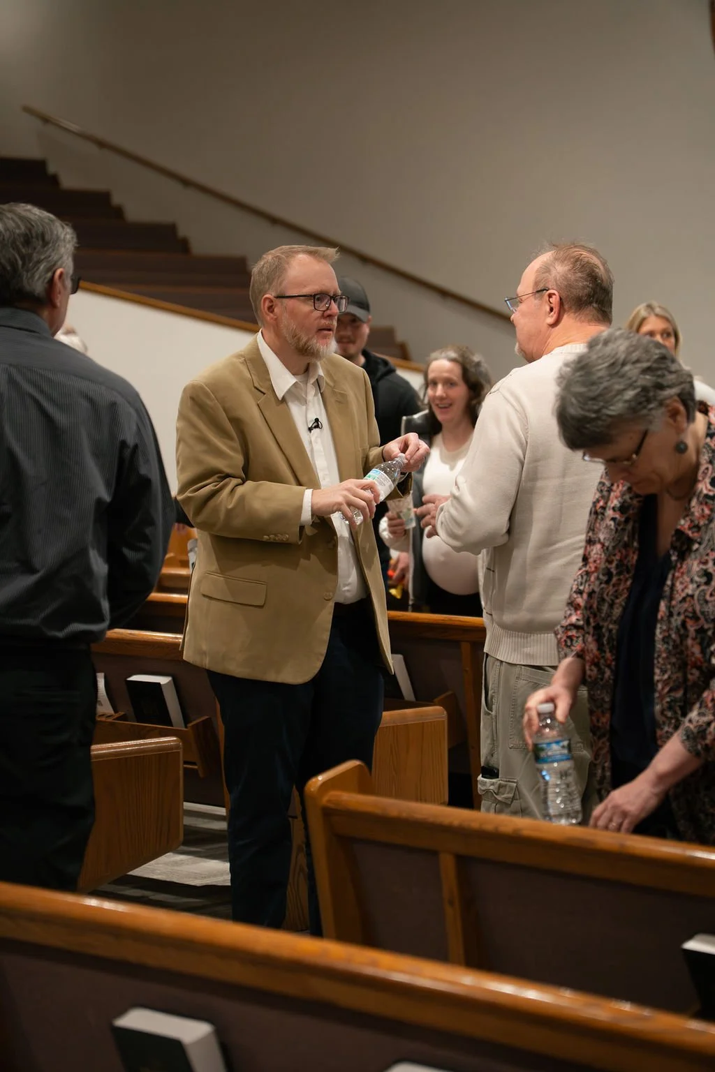Group of people engaging in conversation inside a church or auditorium, some holding water bottles, with wooden pews and a staircase in the background.