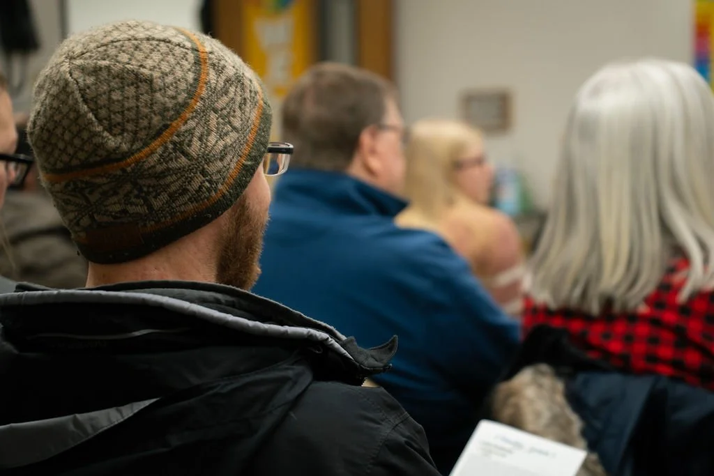 People seated in a room, attending a meeting or presentation, with one man wearing a patterned knit cap in the foreground.