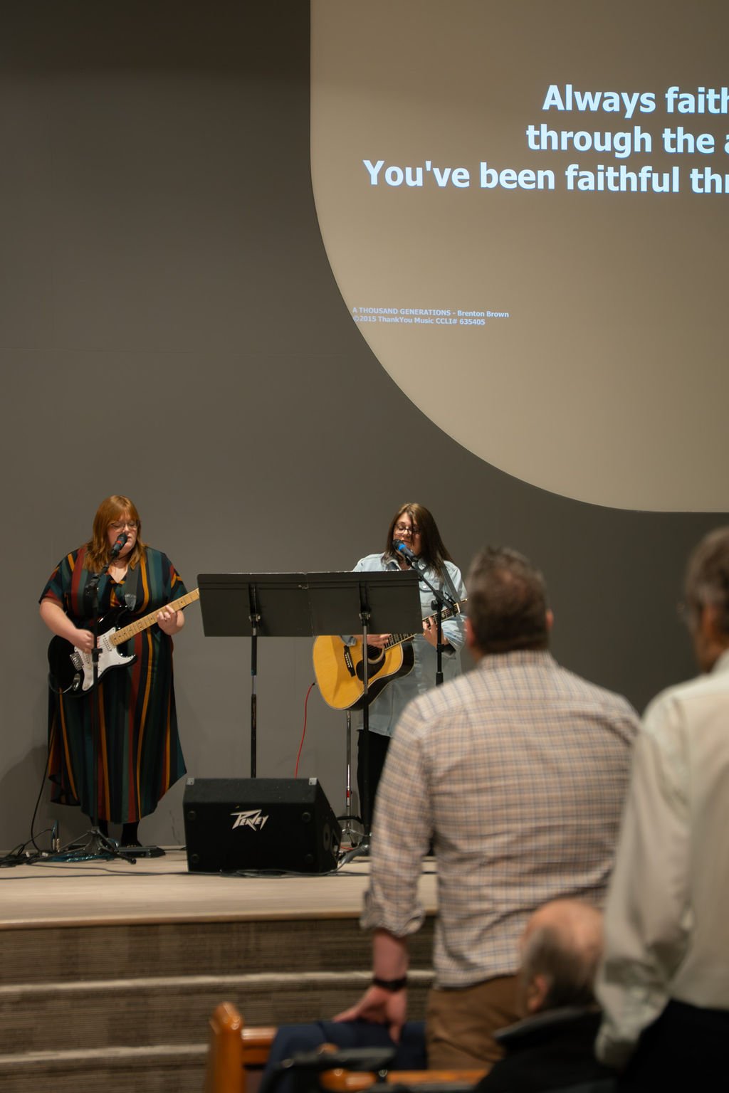 Two women playing guitars and singing on stage, with a large screen behind them displaying song lyrics and people in the audience watching.