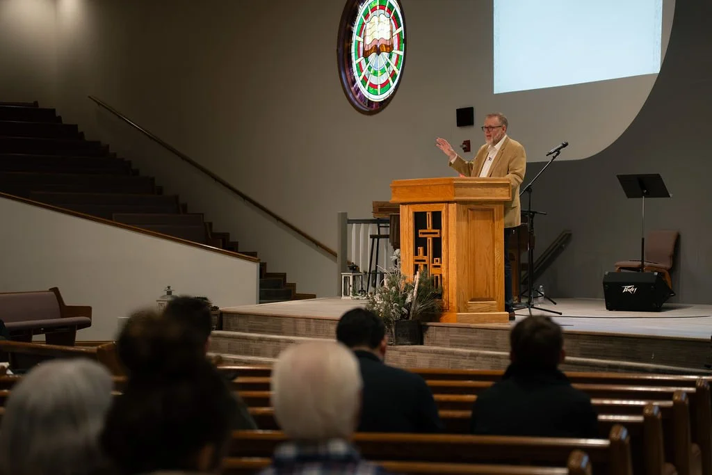 A preacher or speaker standing at a wooden pulpit with a cross design, addressing an audience in a church or chapel. There is a stained glass window, a large projected screen, and musical equipment on the stage.