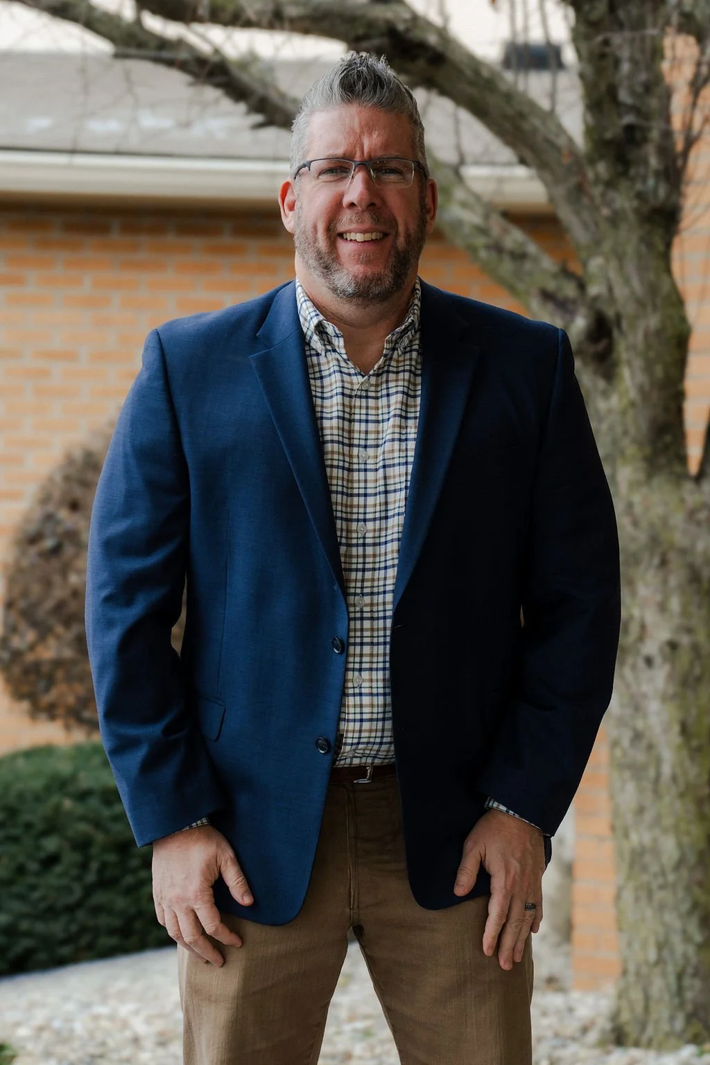 A man with gray hair and glasses standing outdoors, dressed in a blue blazer, plaid shirt, and khaki pants, smiling at the camera.