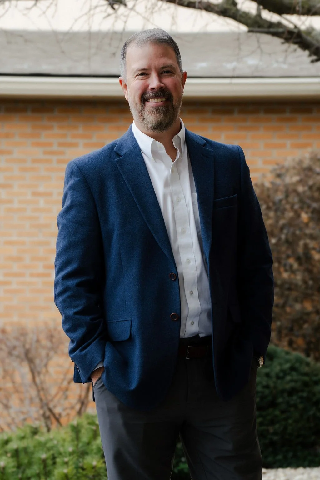 A smiling man in a blue blazer and white shirt standing outside in front of a brick wall and trees.