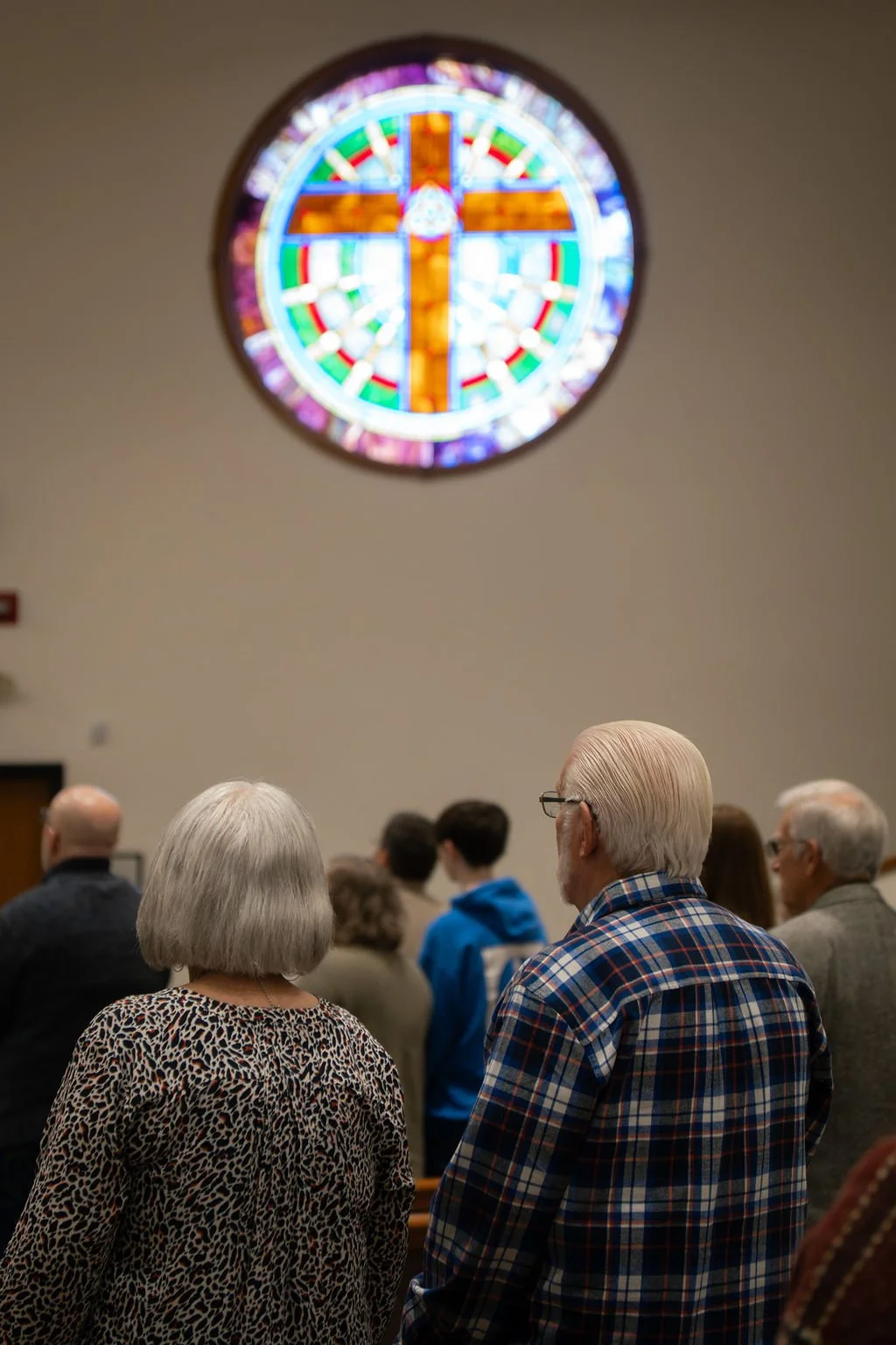 People attending a church service, standing in pews, facing an altar outside the frame, with a large colorful stained glass cross window on the wall above them.