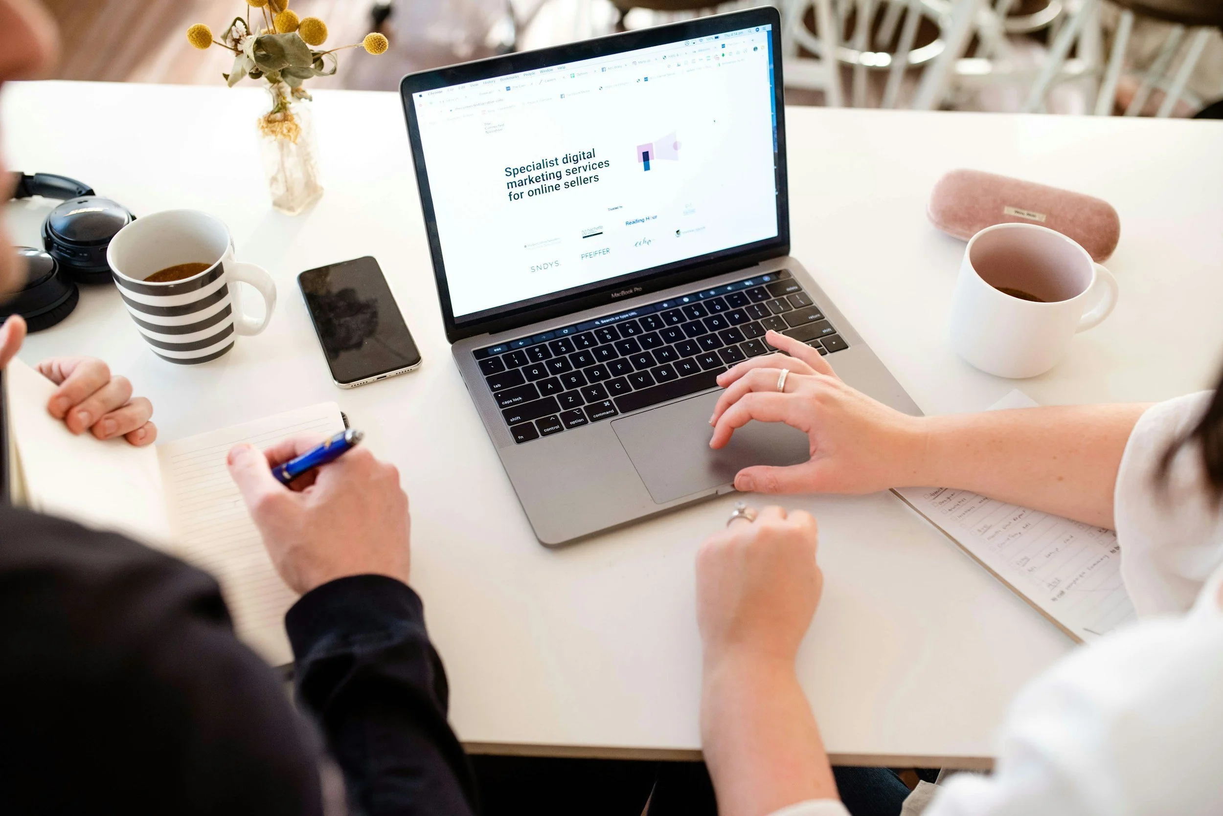 Two people working at a white table with laptops, coffee cups, a smartphone, headphones, a notepad, pens, and a small flower arrangement. One laptop screen displays marketing services for online sellers.