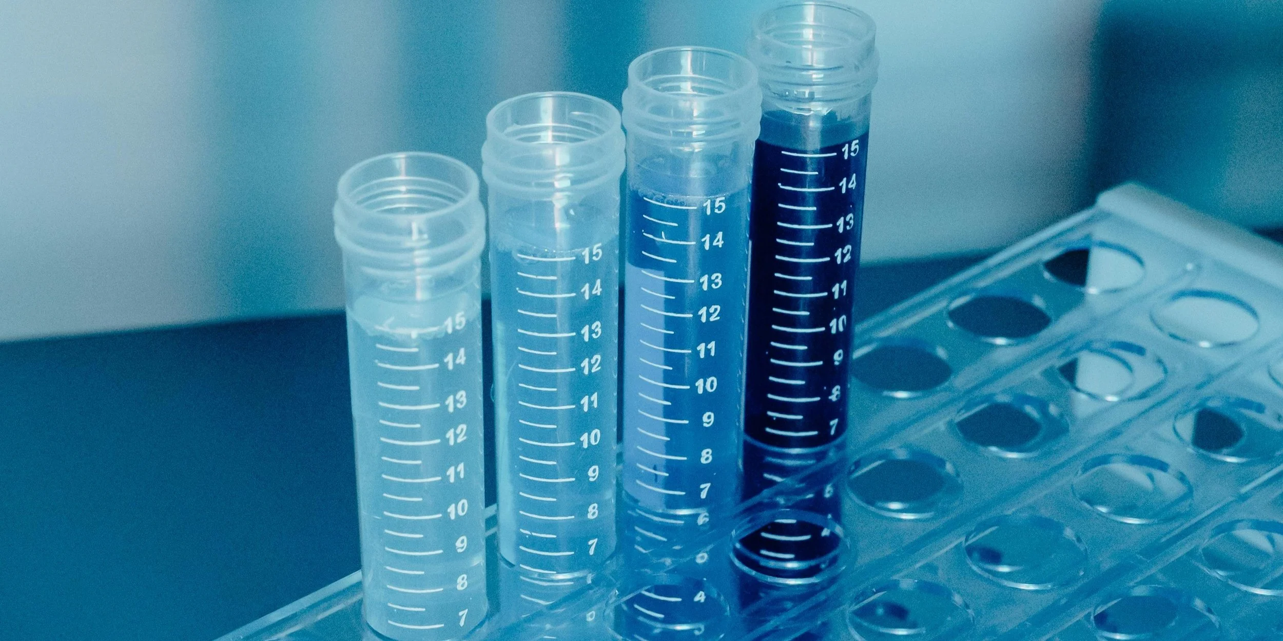 Six clear plastic test tubes with white measurement markings, one filled with dark liquid, placed on a rack in a laboratory setting.