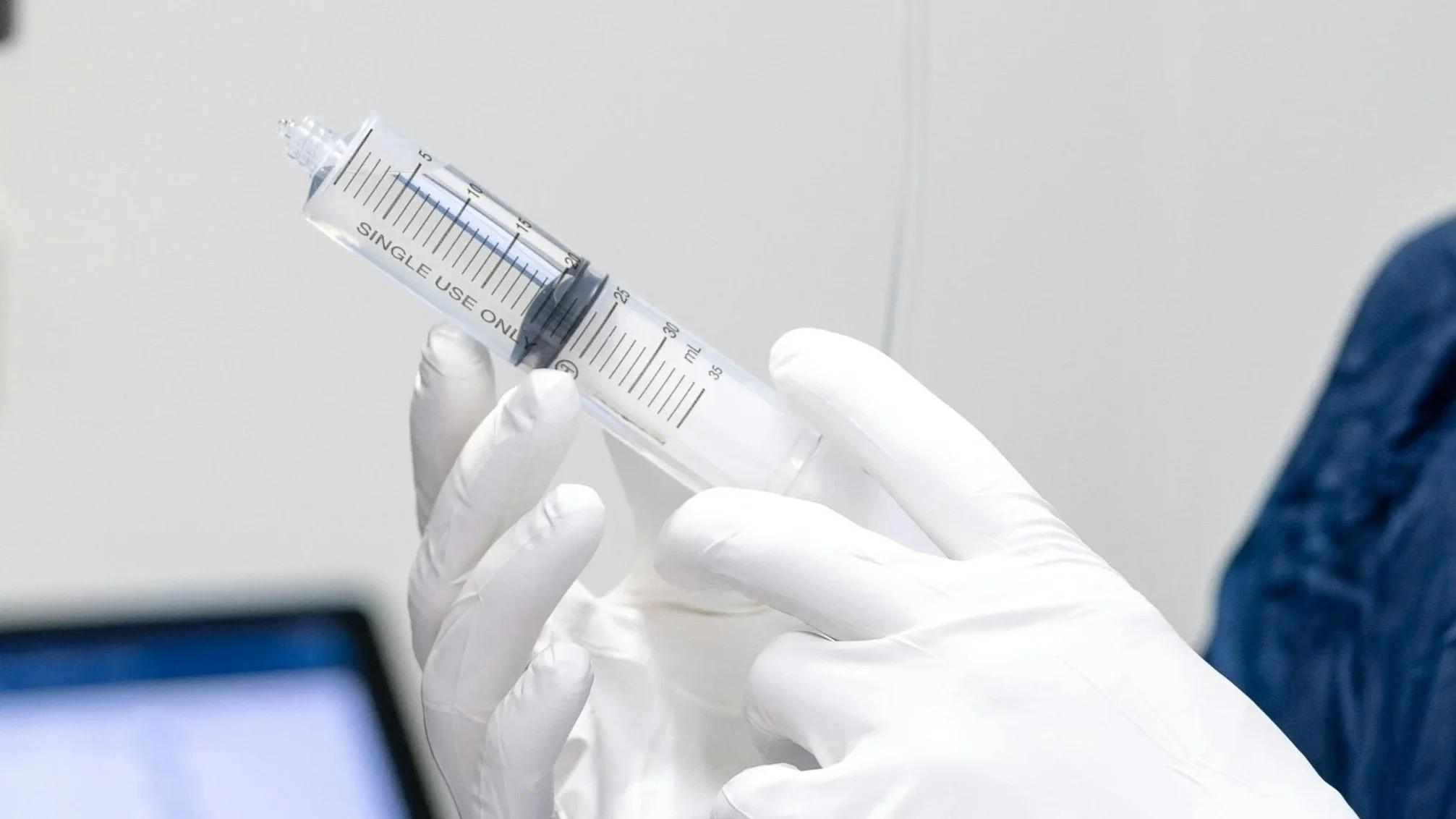 A person wearing white gloves holding a large medical syringe with measurement markings, pointing upwards.