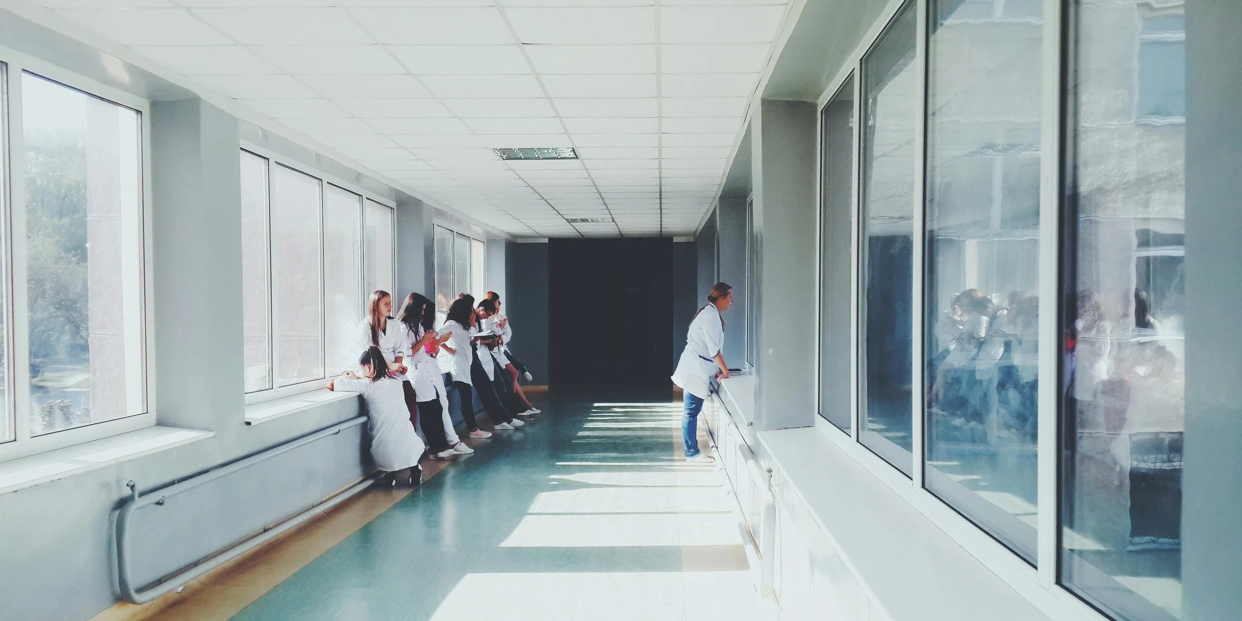 A group of medical students dressed in white coats waiting outside a hospital room, some sitting on the window sill, while a woman in a white coat is standing near the window viewing inside.