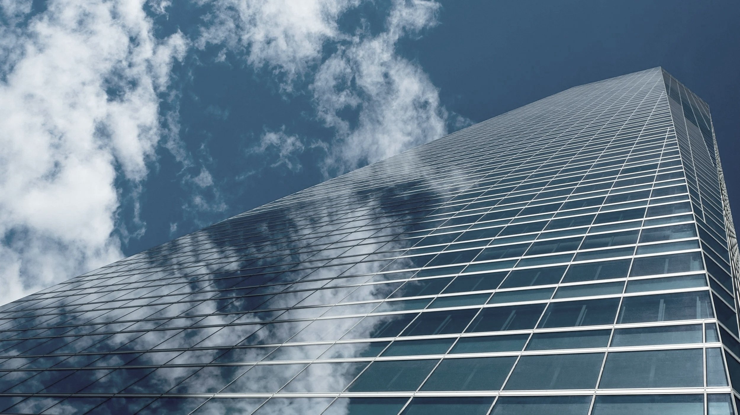 Looking up at a modern glass skyscraper with reflections of clouds and the blue sky.