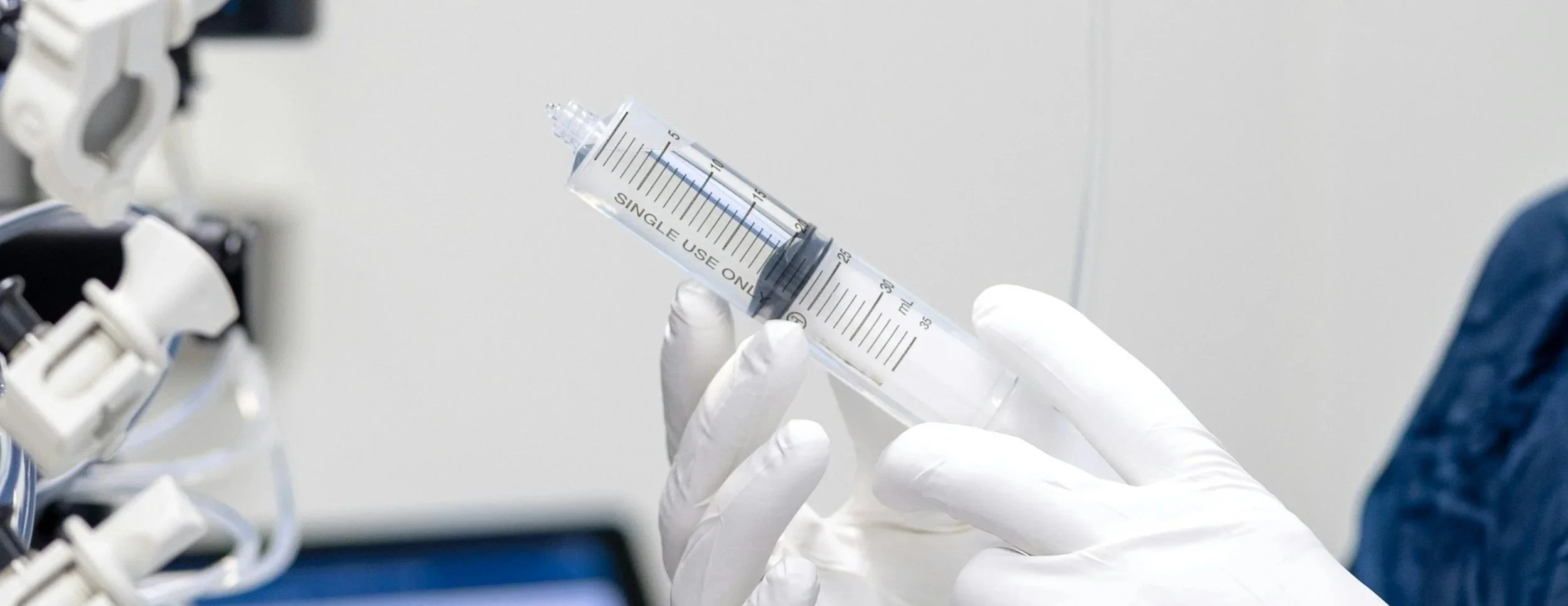 A medical professional wearing white gloves holding a large syringe inside a clinical setting.