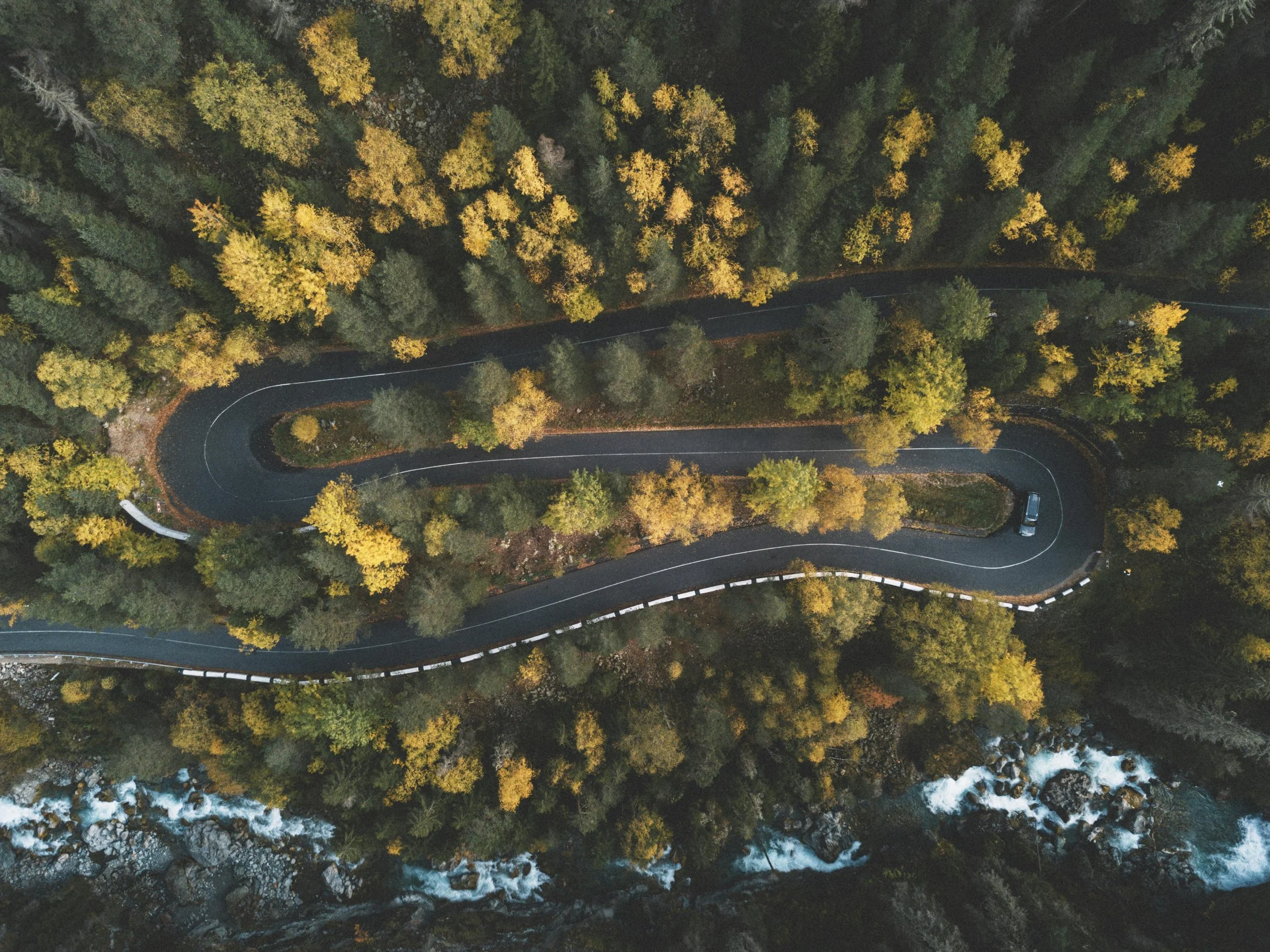An aerial view of a winding mountain road through a forest with yellow and green trees, near a rocky river with white water at the bottom.