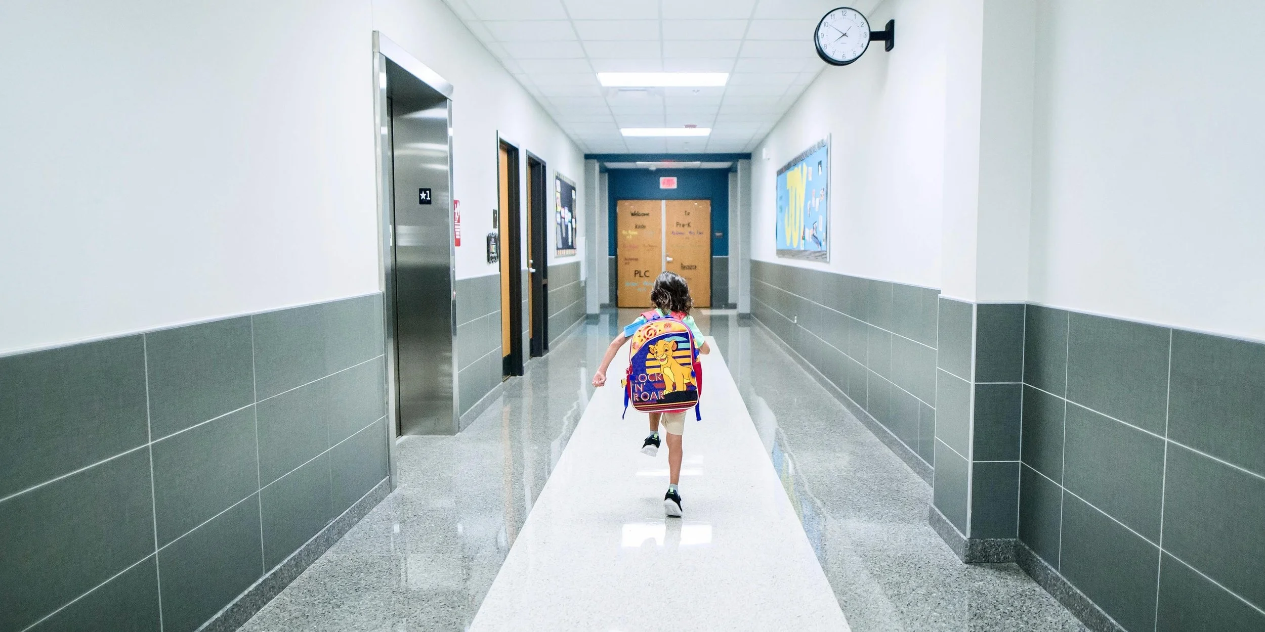 A young girl with a colorful backpack running down a school hallway.