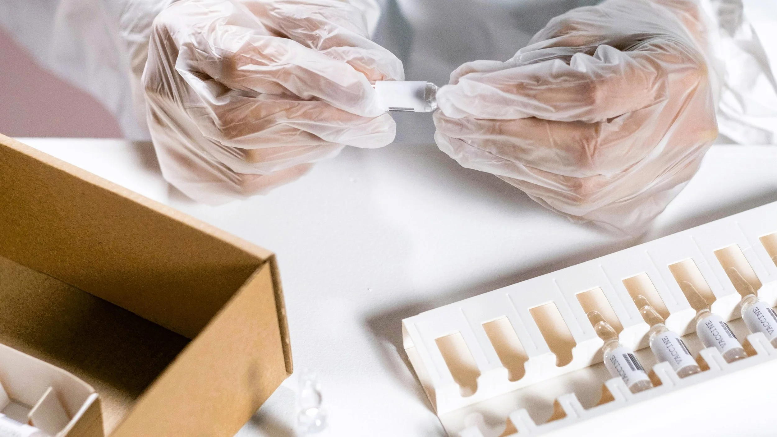 Person wearing white gloves filling a syringe with medication or vaccine from a vial, with medical supplies and a cardboard box on a white surface.