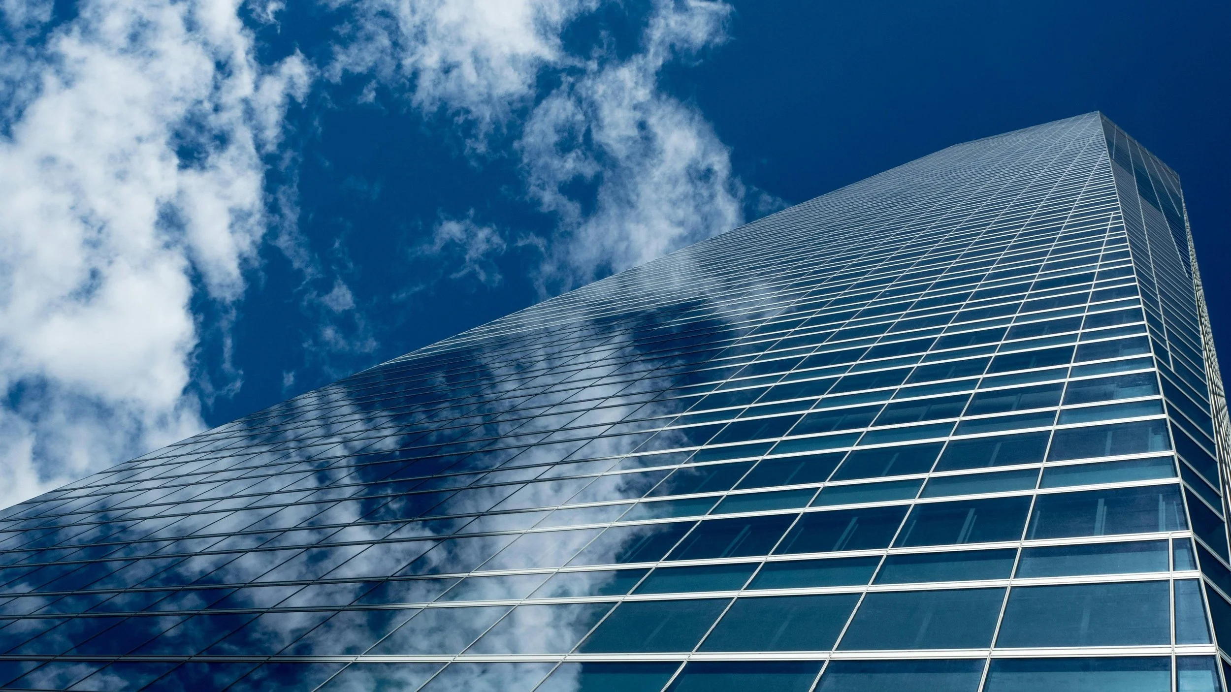 A tall glass skyscraper reflecting the blue sky and white clouds on its mirrored surface