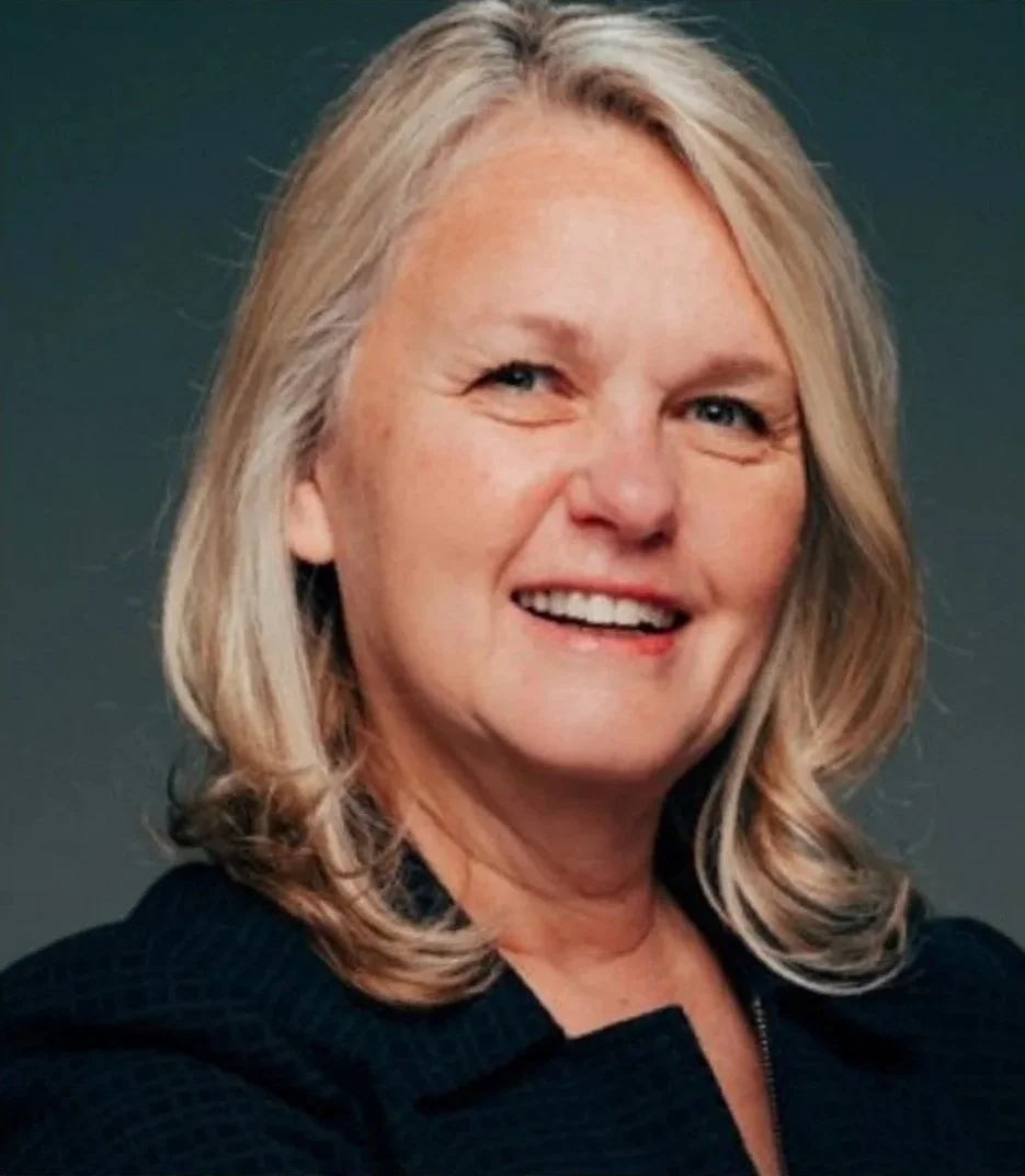 A portrait of a smiling middle-aged woman with shoulder-length blonde hair, wearing a dark top, set against a dark background.