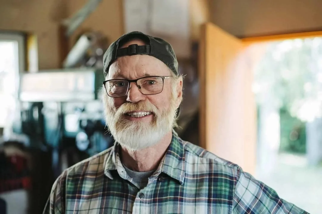 Smiling elderly man with a beard, wearing glasses, a backwards cap, and a plaid shirt inside a cozy wood-paneled room.