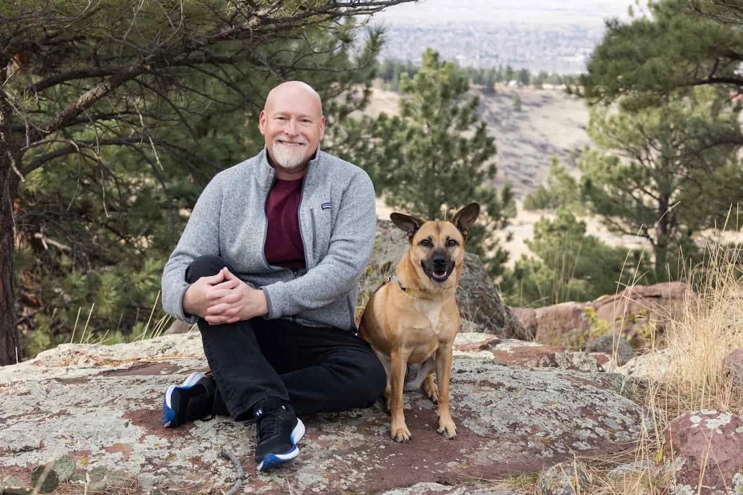 A smiling man with a beard and a dog sitting on a large rock in a forested mountainous area.