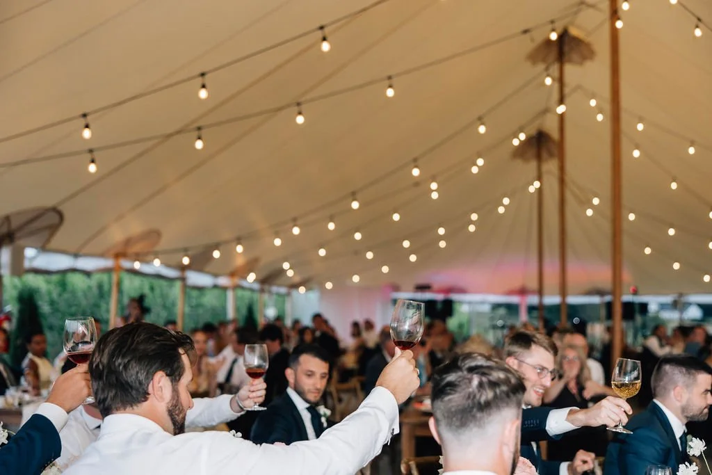 Guests at a celebration raising glasses of wine under a large tent decorated with string lights.