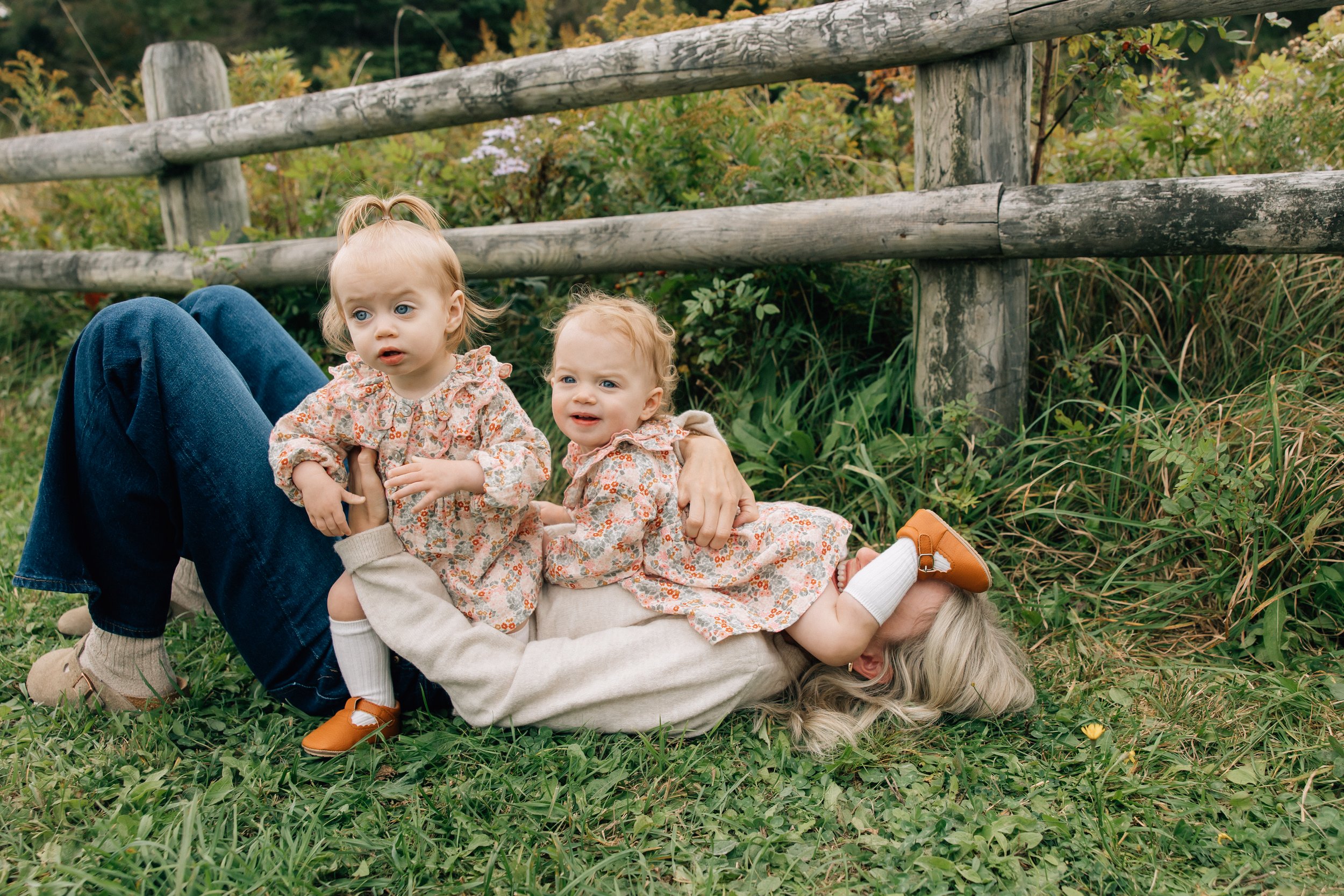 A woman is lying on the grass outdoors with two young girls, all dressed in casual clothing, surrounded by green grass and plants, near a wooden fence.