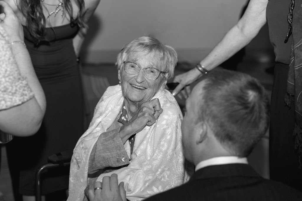 Older woman with glasses smiling, surrounded by people at a social gathering.