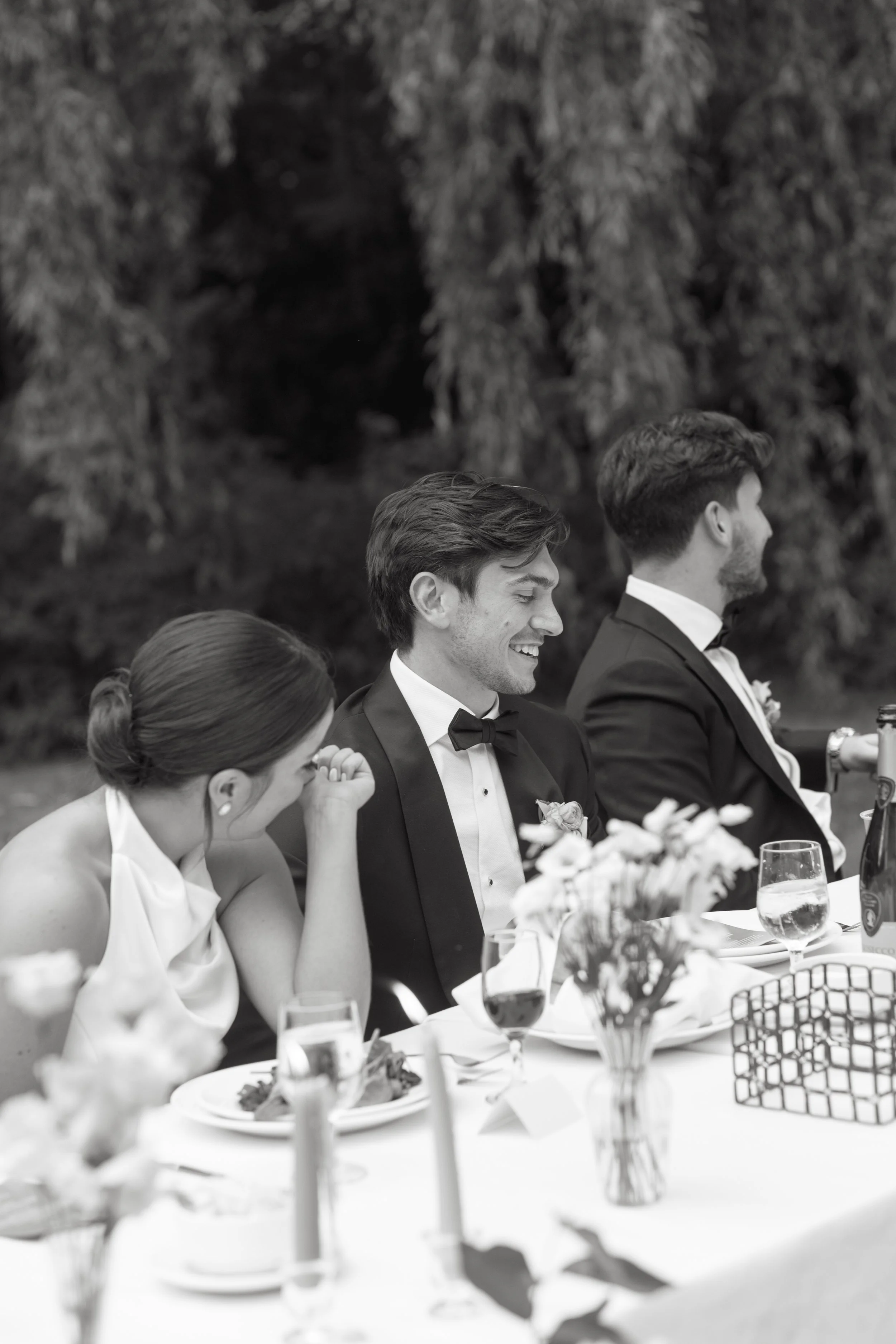 Black and white photo of a wedding reception with three well-dressed people sitting at a table, two men in tuxedos and a woman in a dress, with food, drinks, and flowers on the table, outdoors with trees in the background.