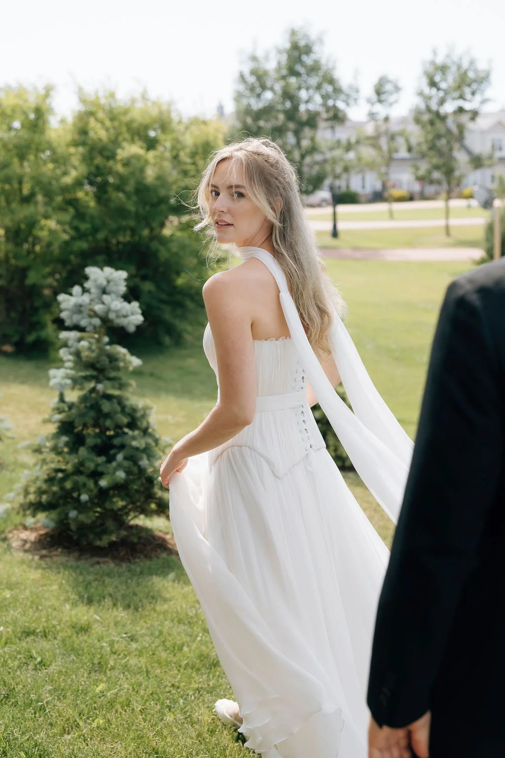 A young woman in a white dress standing outdoors in a garden, looking back over her shoulder, with greenery and trees in the background.
