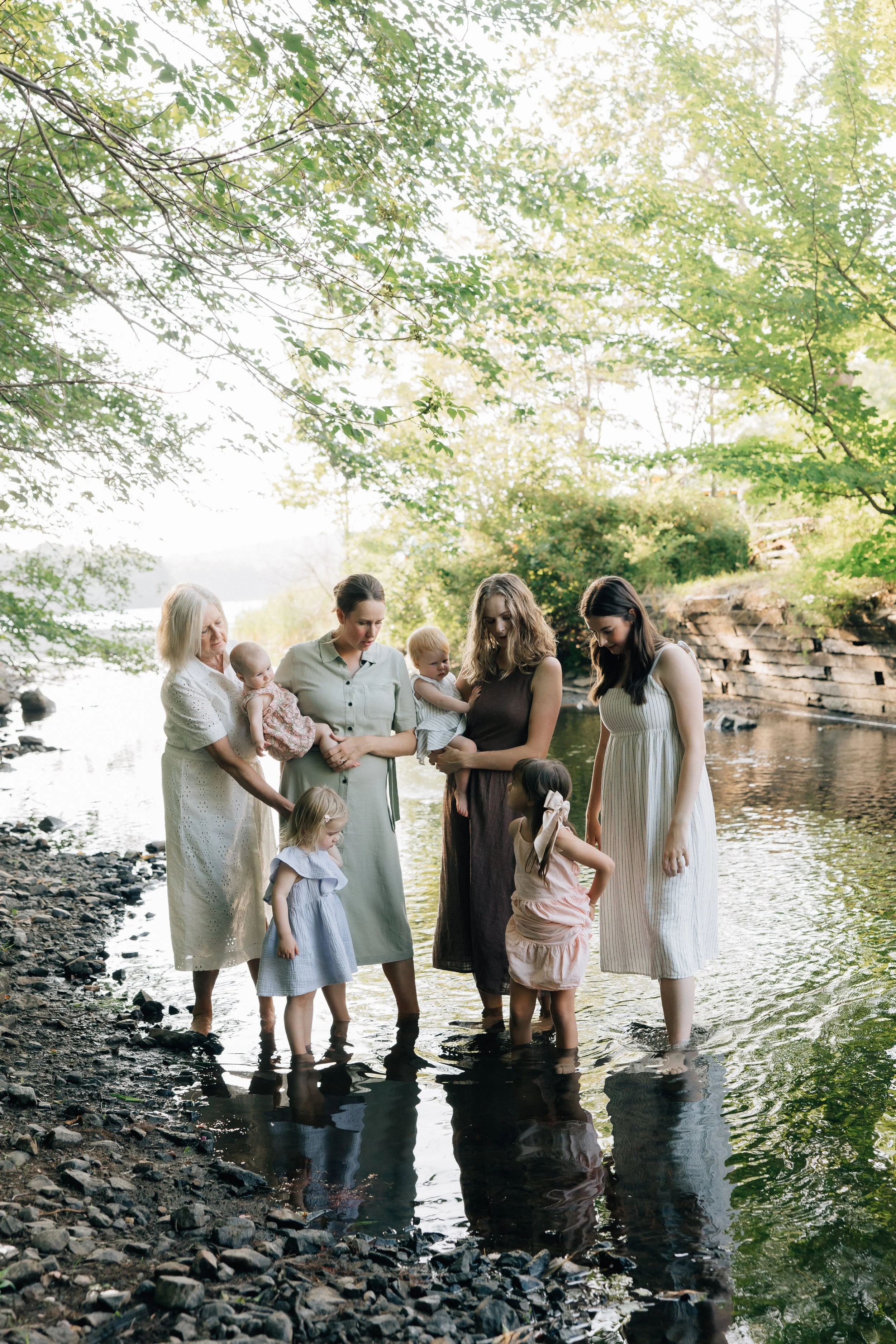 A group of women and children standing in a shallow river surrounded by trees with green leaves, holding hands and looking down at the water.
