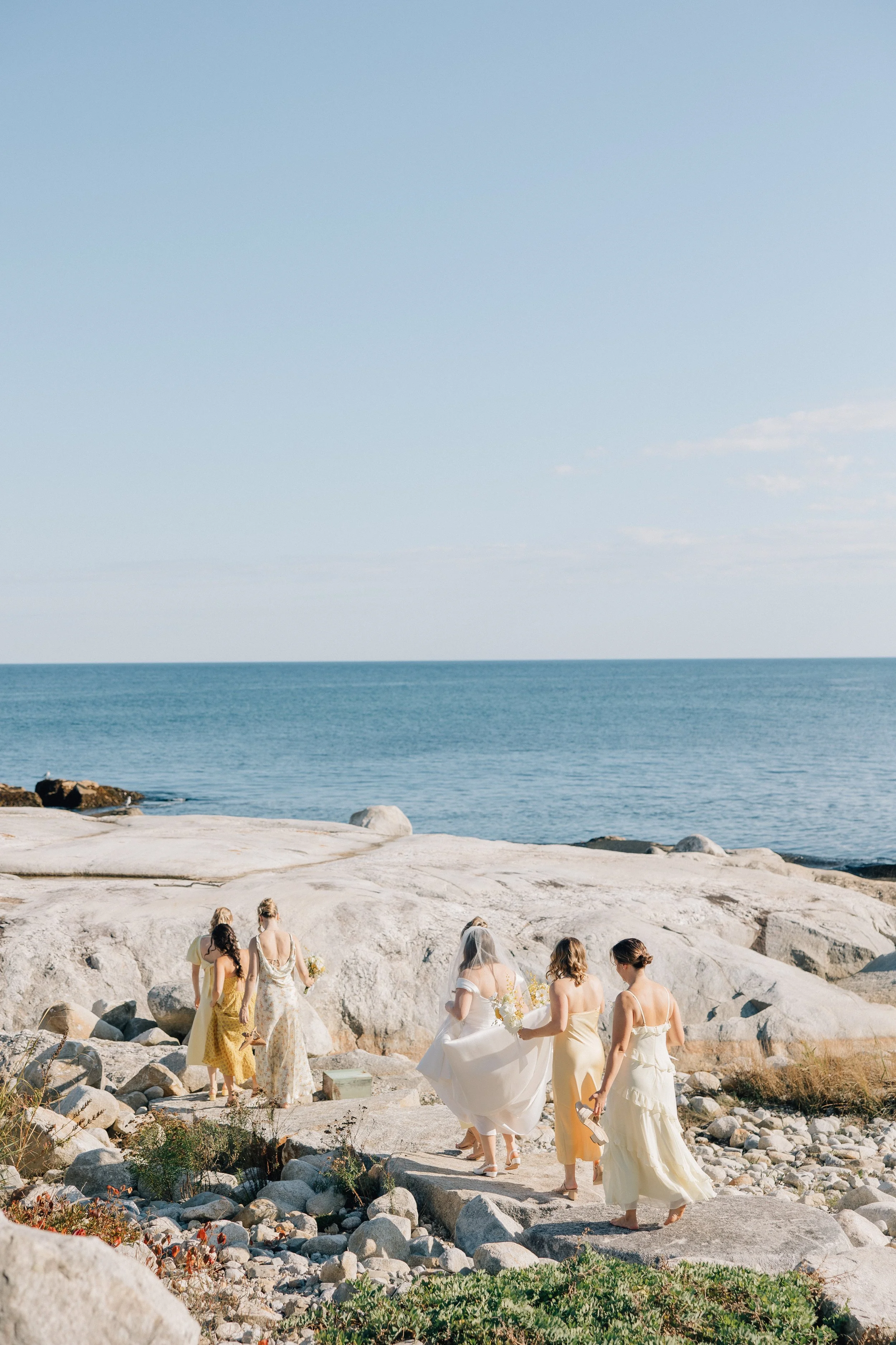 A group of women dressed in white and pastel dresses walking along a rocky beach, possibly for a wedding, with the ocean and a clear sky in the background.
