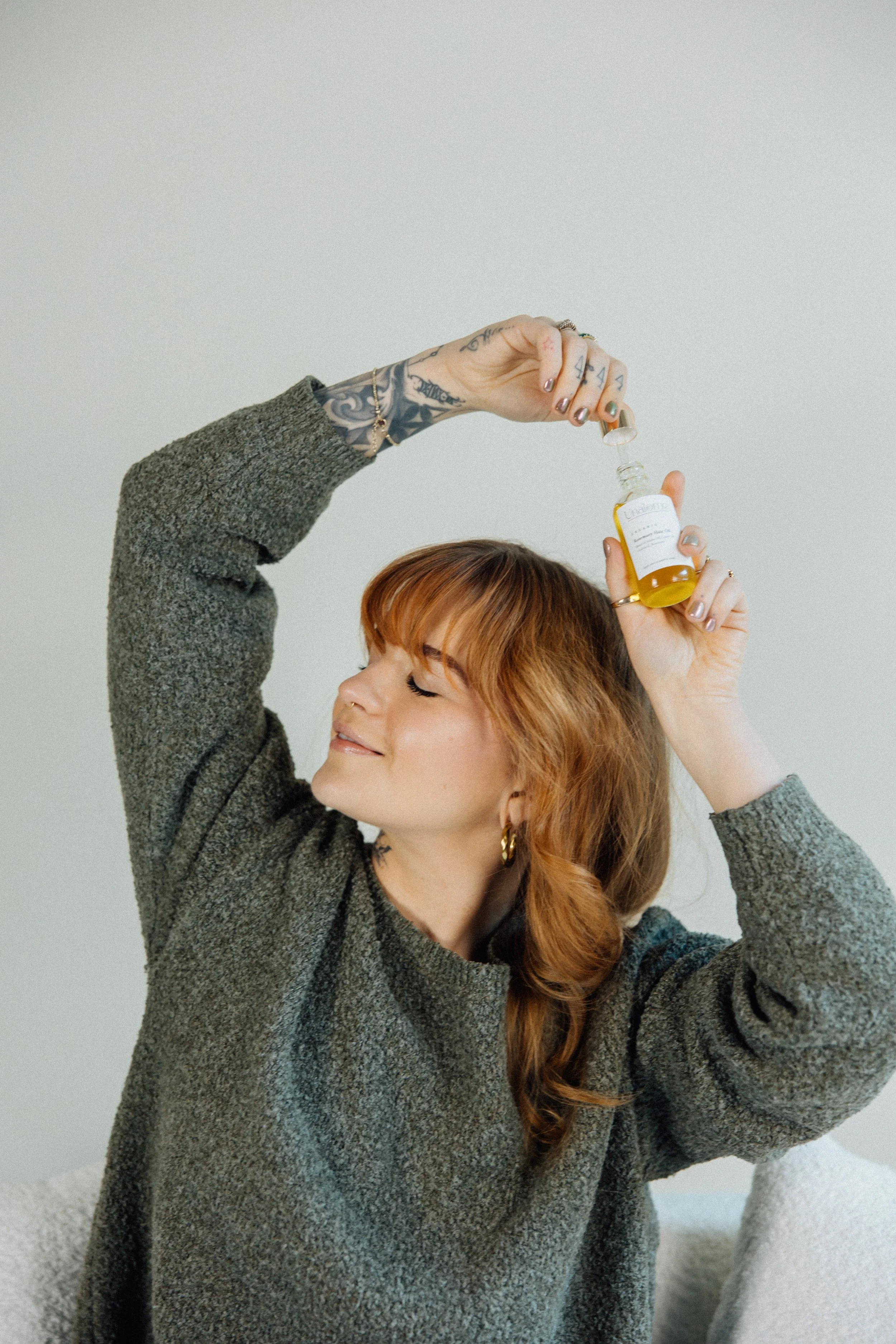 A young woman with red hair and tattoos on her right hand and left arm is smiling with her eyes closed, holding a small dropper bottle with a yellow liquid. She is wearing a gray knit sweater and standing in front of a plain light gray wall.
