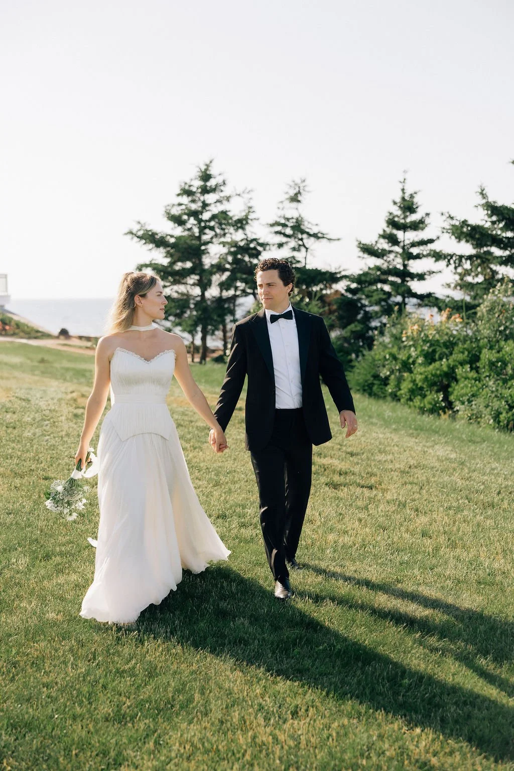 A bride and groom walking hand in hand on a grassy area outdoors, with trees and the ocean in the background, during their wedding day.