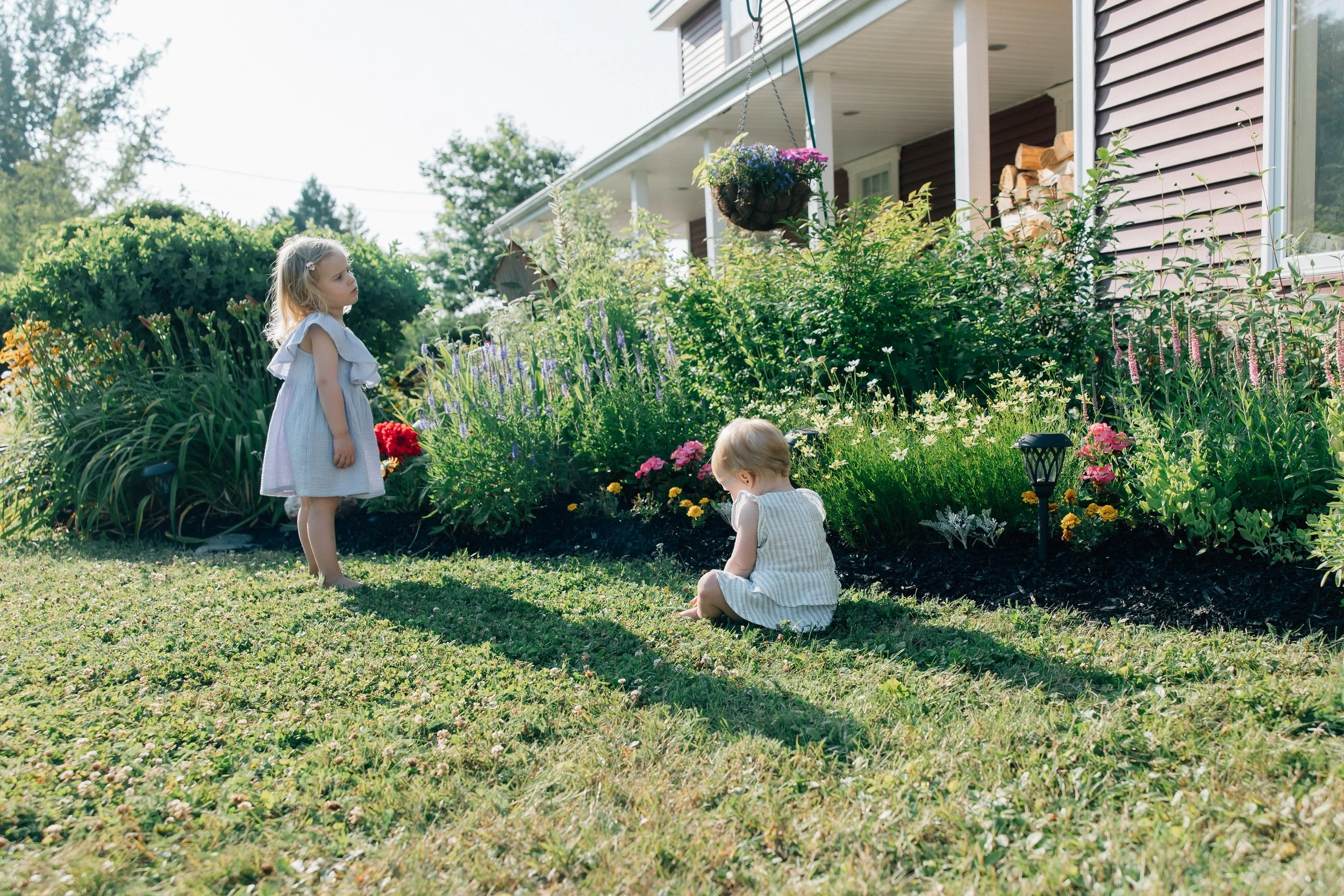 Two young girls playing in a garden with colorful flowers, a hanging flower pot, and a house with wood logs in the background on a sunny day.
