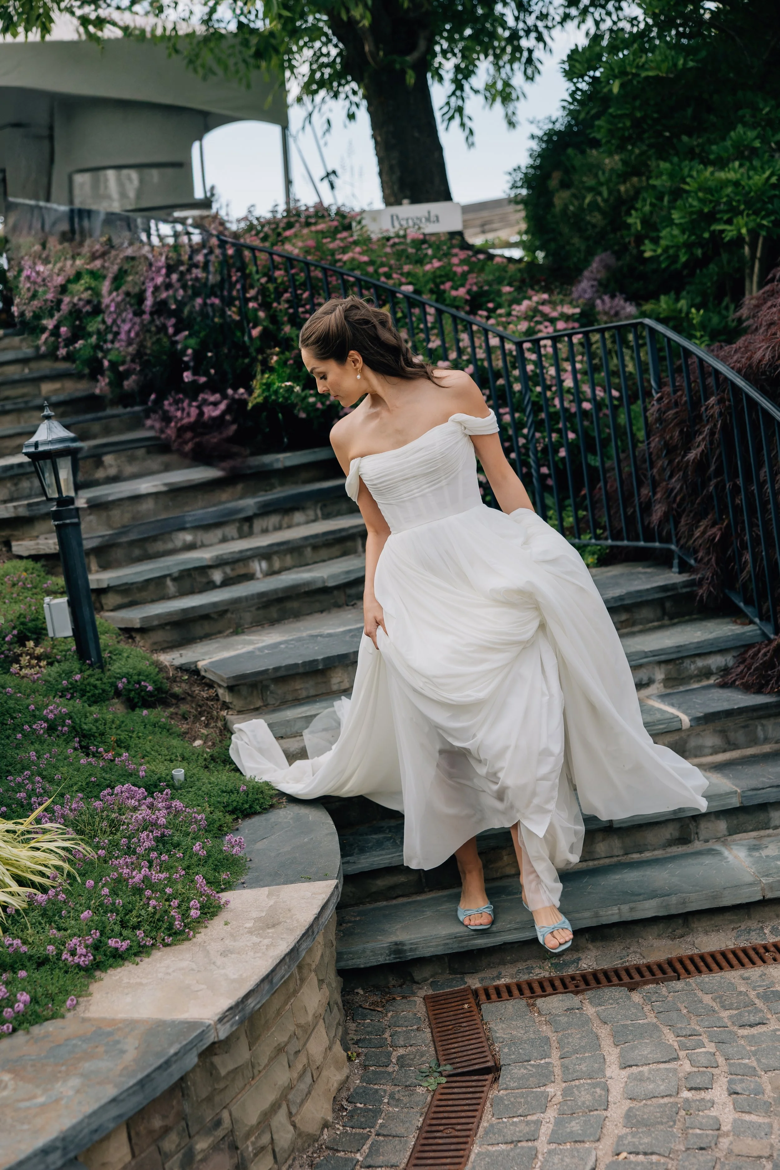 A woman in a white off-shoulder dress walking down stone stairs surrounded by pink and purple flowers and green bushes.